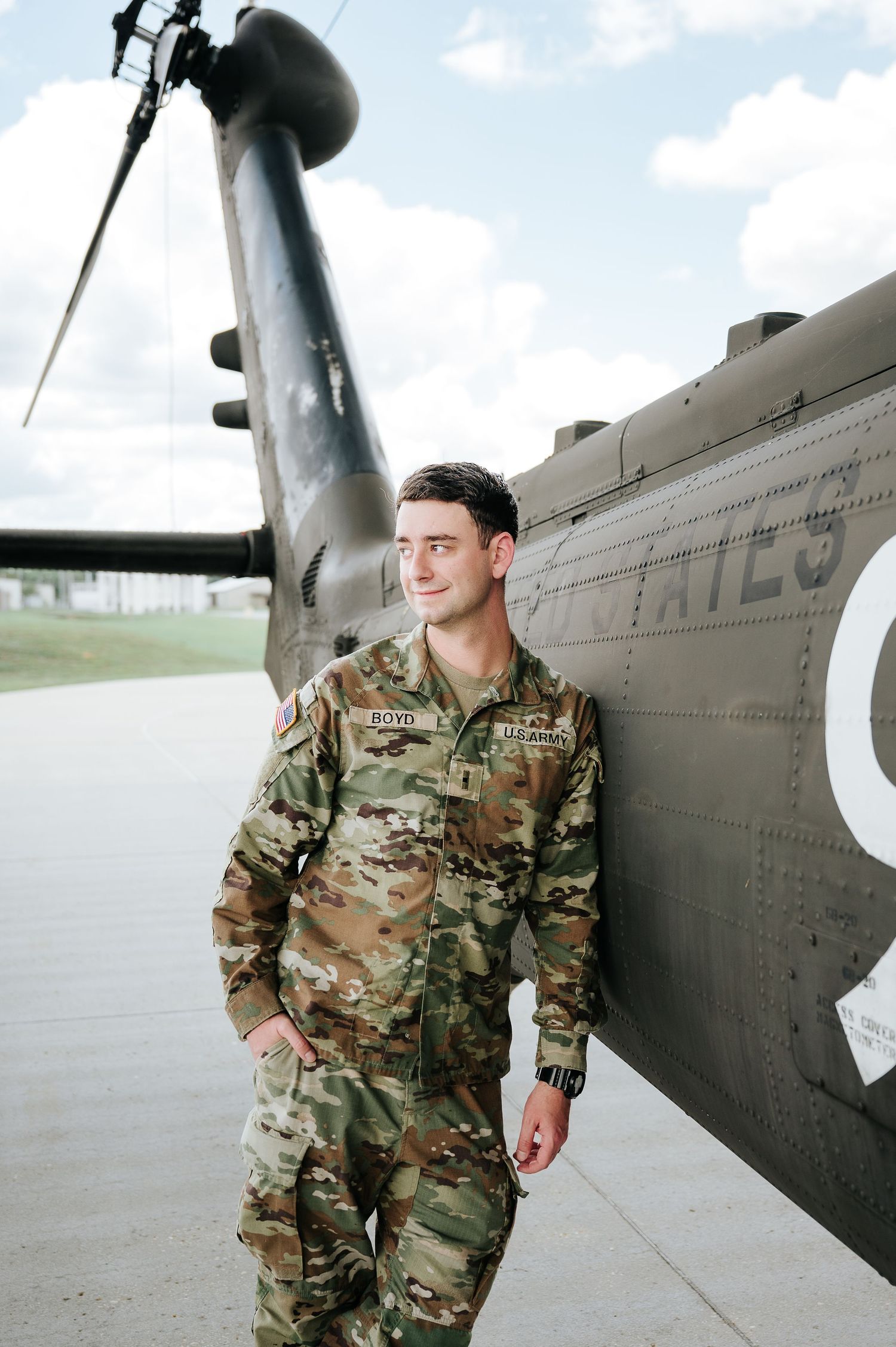 Military soldier in camouflage uniform leaning against a helicopter on an airfield, smiling confidently.