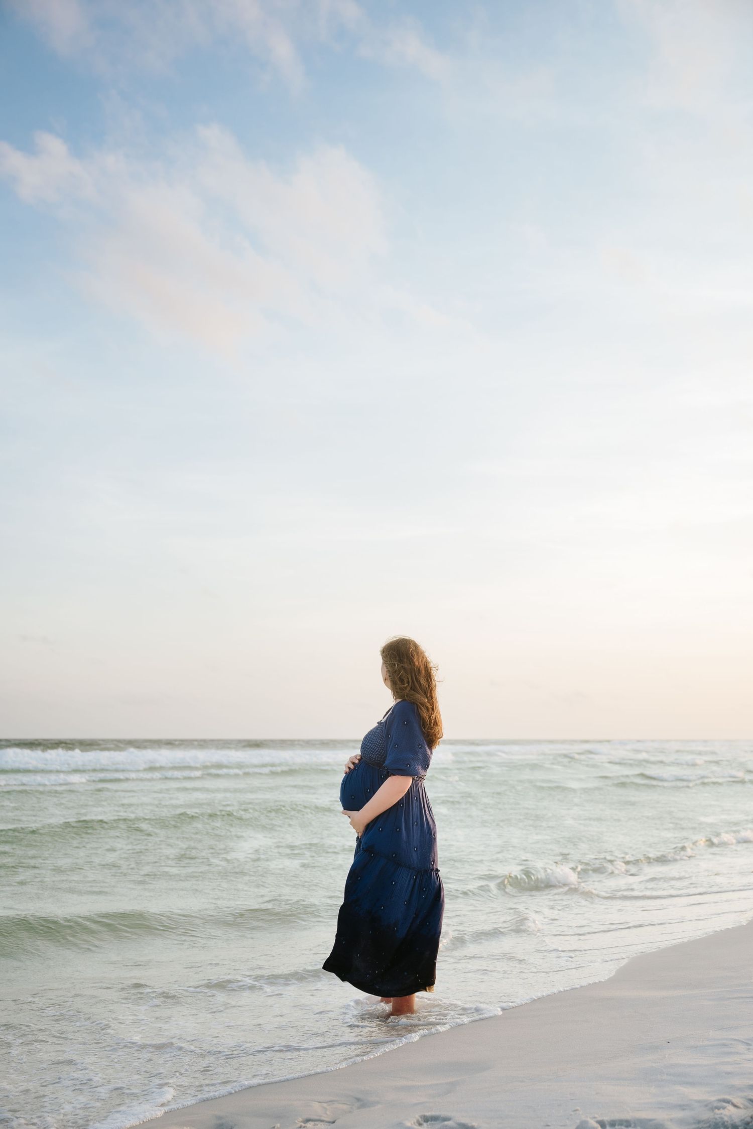 Pregnant woman in navy blue dress standing at ocean shoreline during sunset, cradling her baby bump.