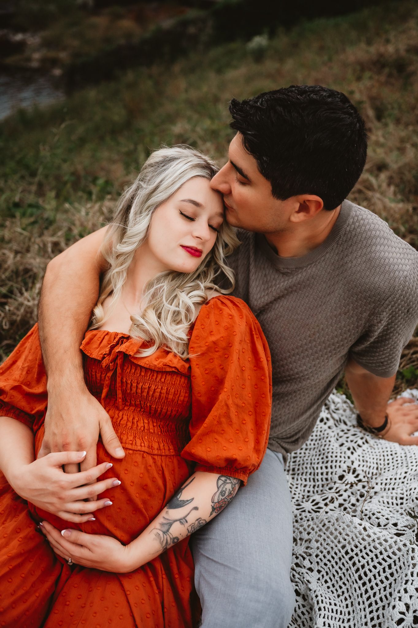A romantic maternity photo of a couple sitting on a blanket outdoors, with the woman wearing a rust-colored dress.