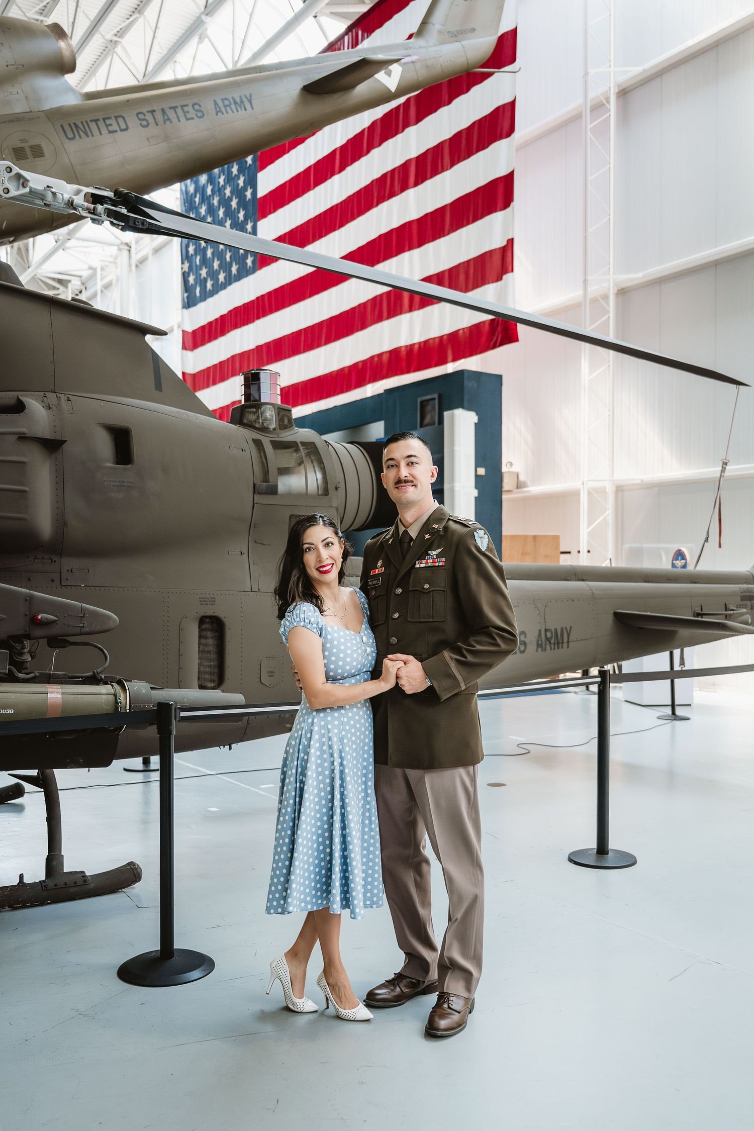 Couple posing in military uniform and blue polka dot dress at an army museum with helicopter and American flag.