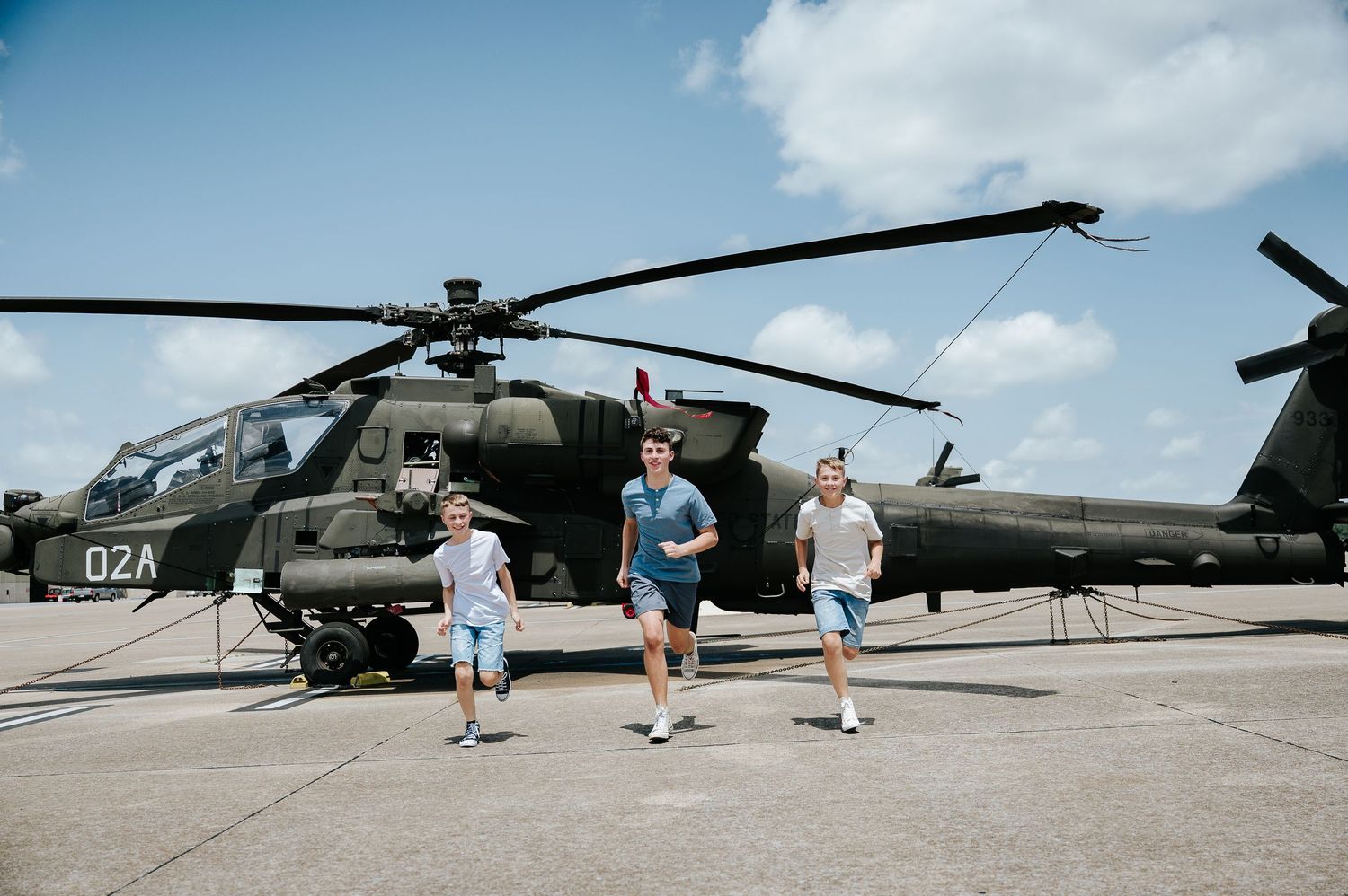 Three young men walk in front of a military Apache helicopter on a sunny airfield tarmac.