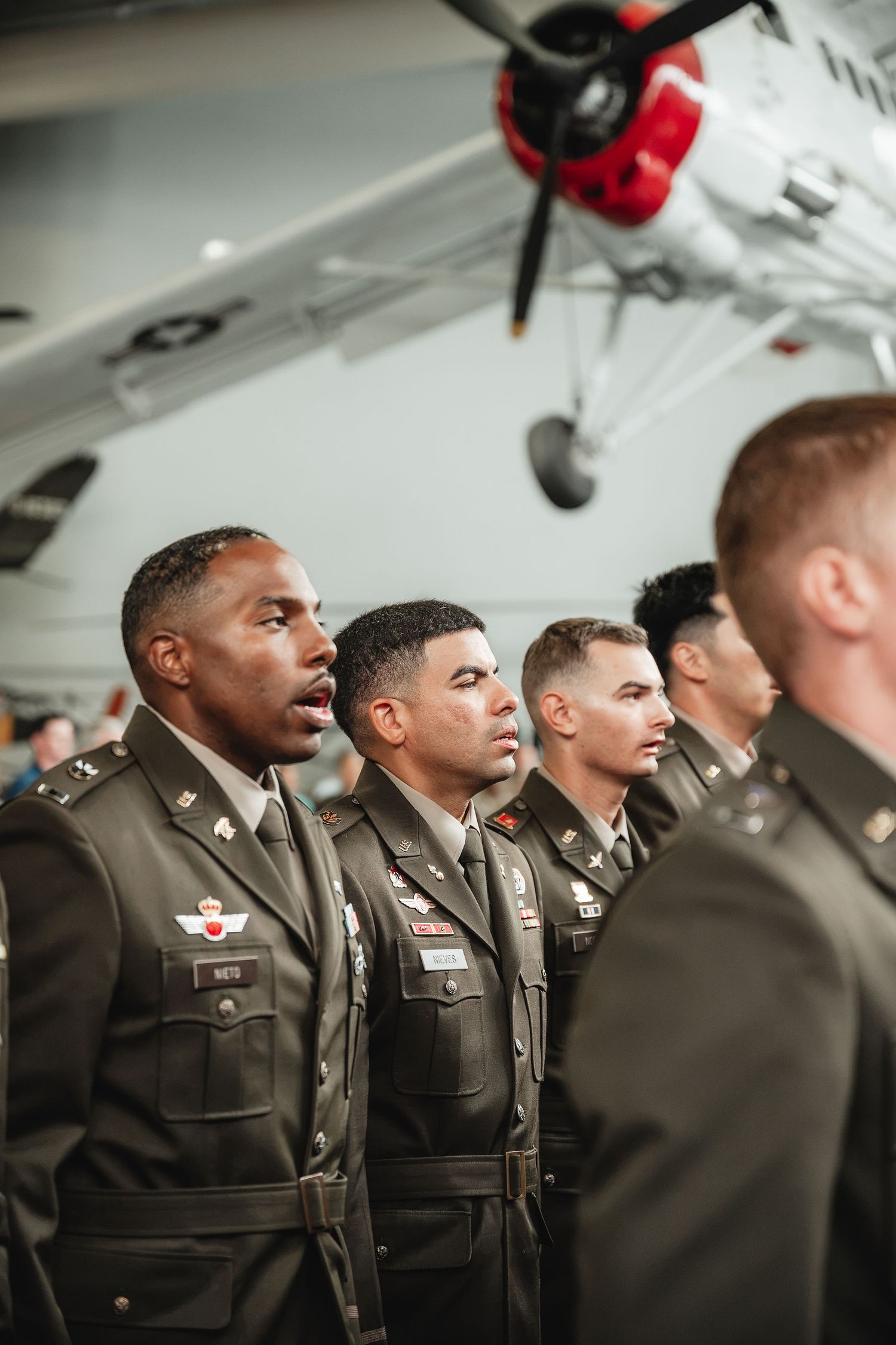 Military officers in dress uniforms stand at attention in a museum hangar with a vintage aircraft displayed overhead.