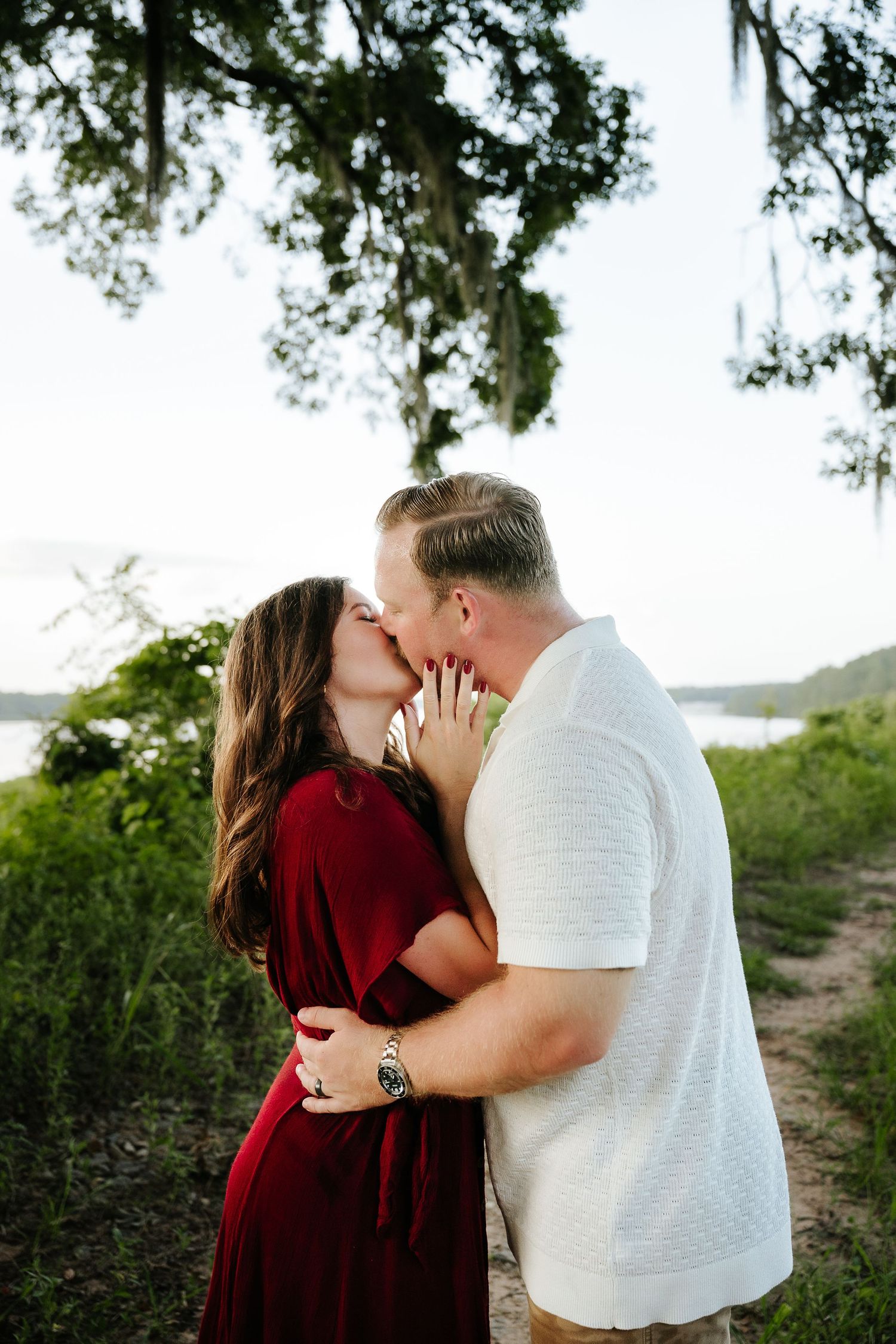 Couple sharing a romantic kiss outdoors near a lake, woman in red dress and man in white shirt embracing among lush greenery.