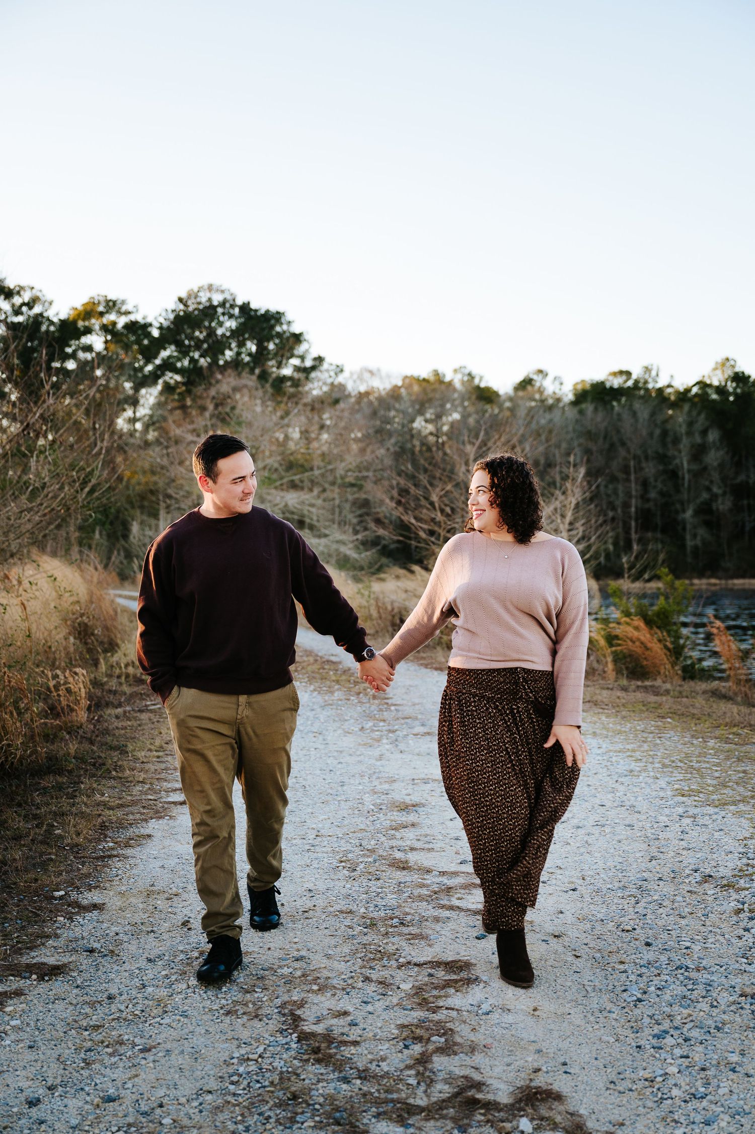 A couple walking together on a gravel path through a wooded area during golden hour.
