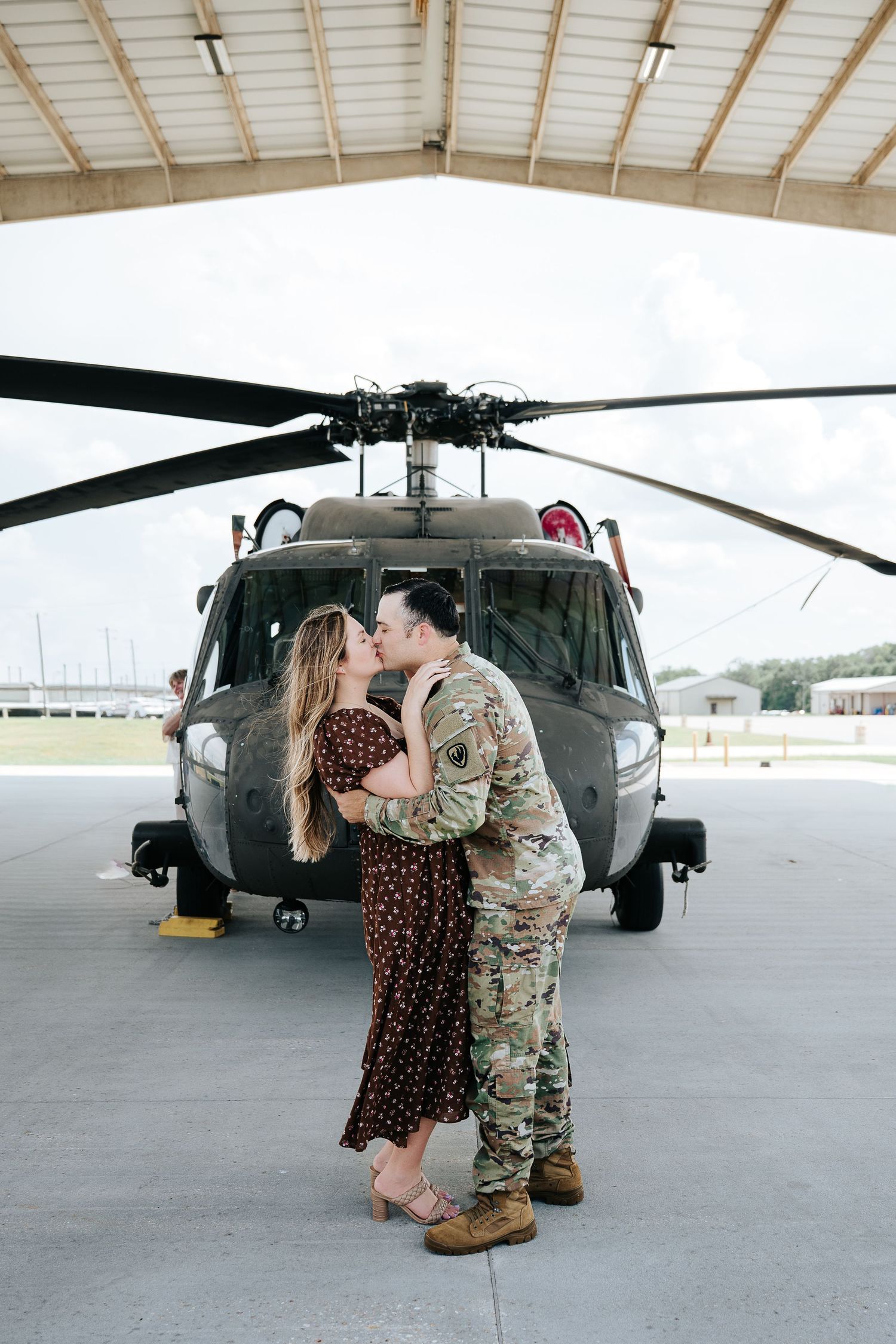 Military couple sharing a romantic kiss in front of a helicopter in a hangar, man in camouflage uniform.