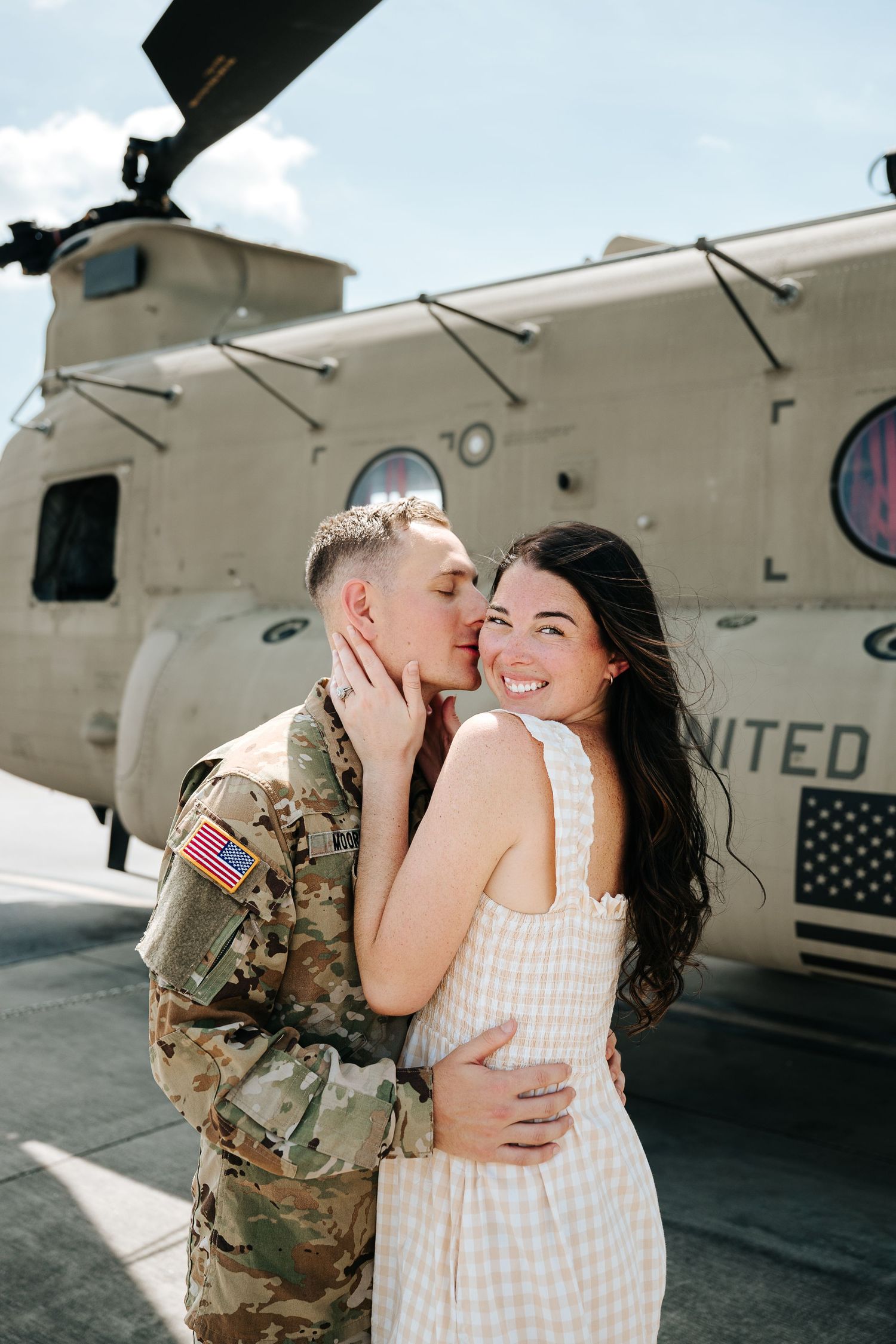 A soldier in camouflage uniform kisses a smiling woman in a pink dress in front of a military helicopter.