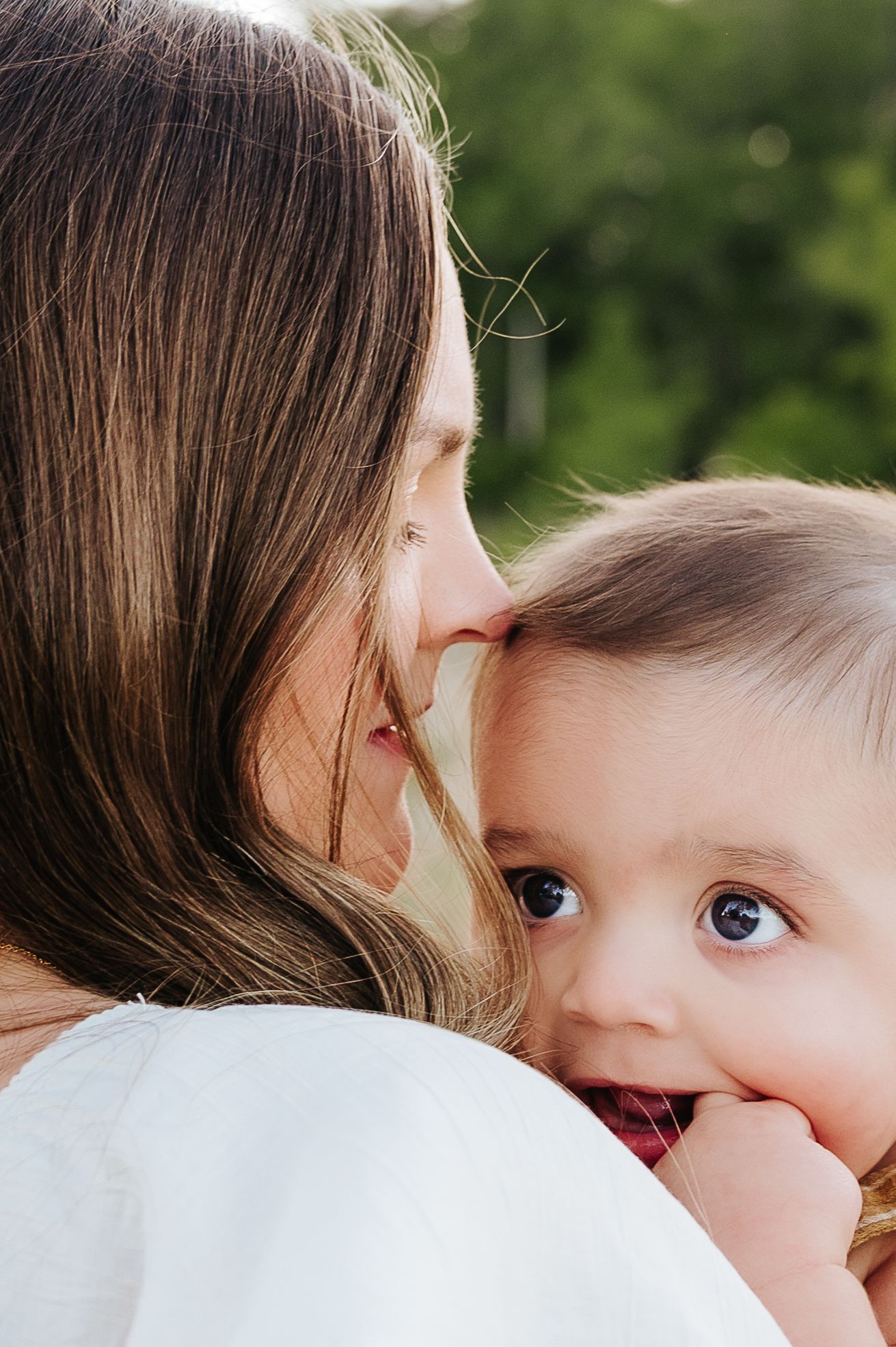 A mother tenderly holds her wide-eyed baby close, sharing an intimate moment outdoors with green foliage behind them.