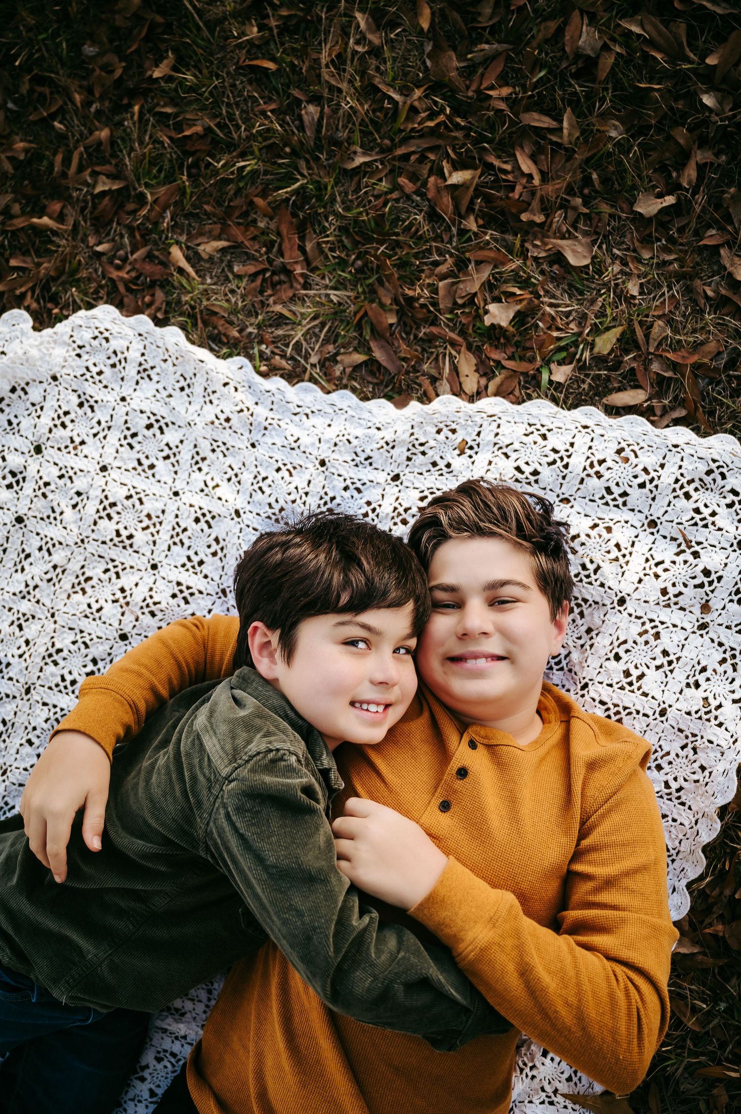 Two boys lying on a white lace blanket outdoors, smiling and hugging surrounded by autumn leaves.