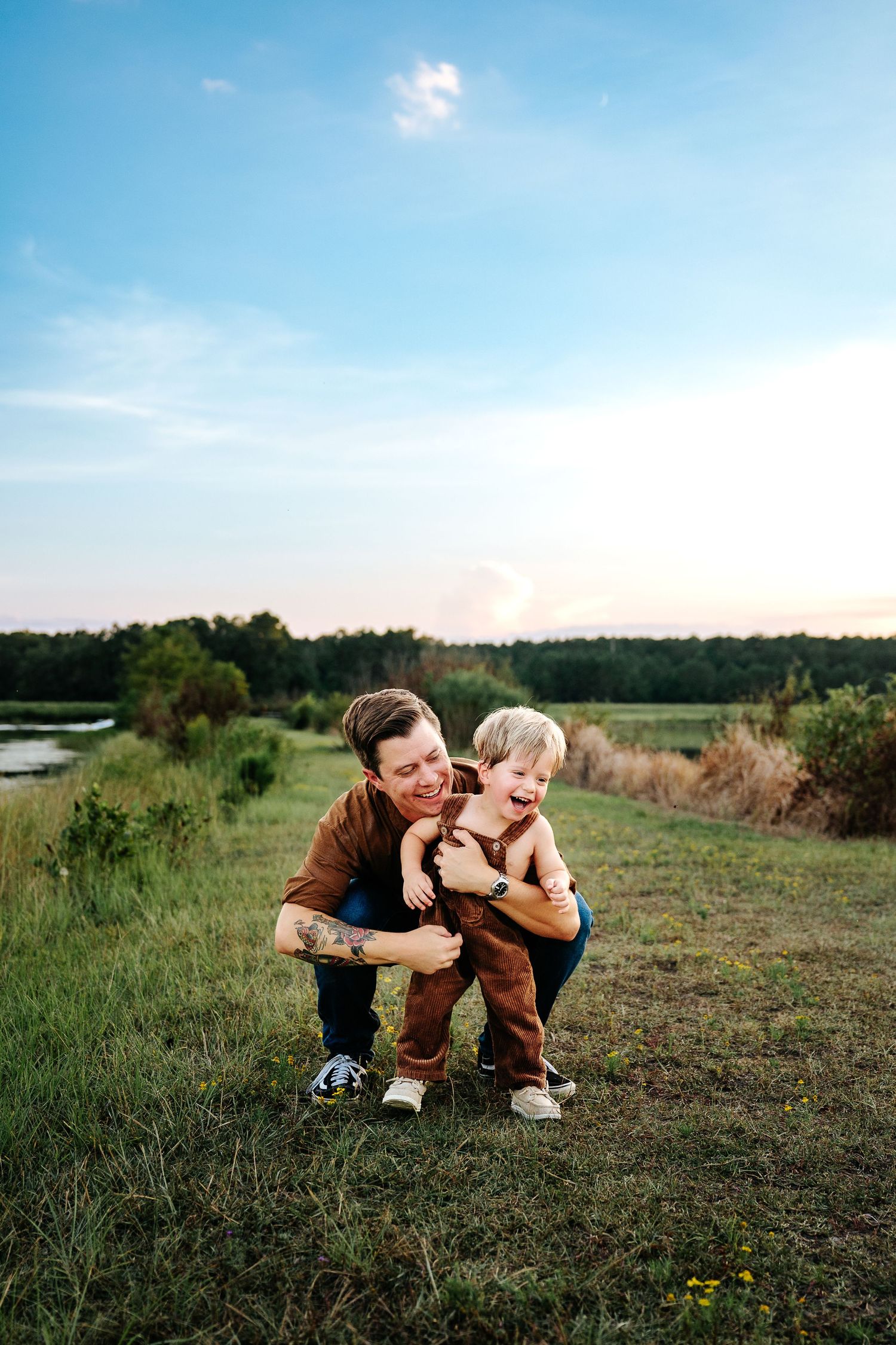 A parent crouches behind a laughing toddler in a green field at sunset, both wearing brown outfits.