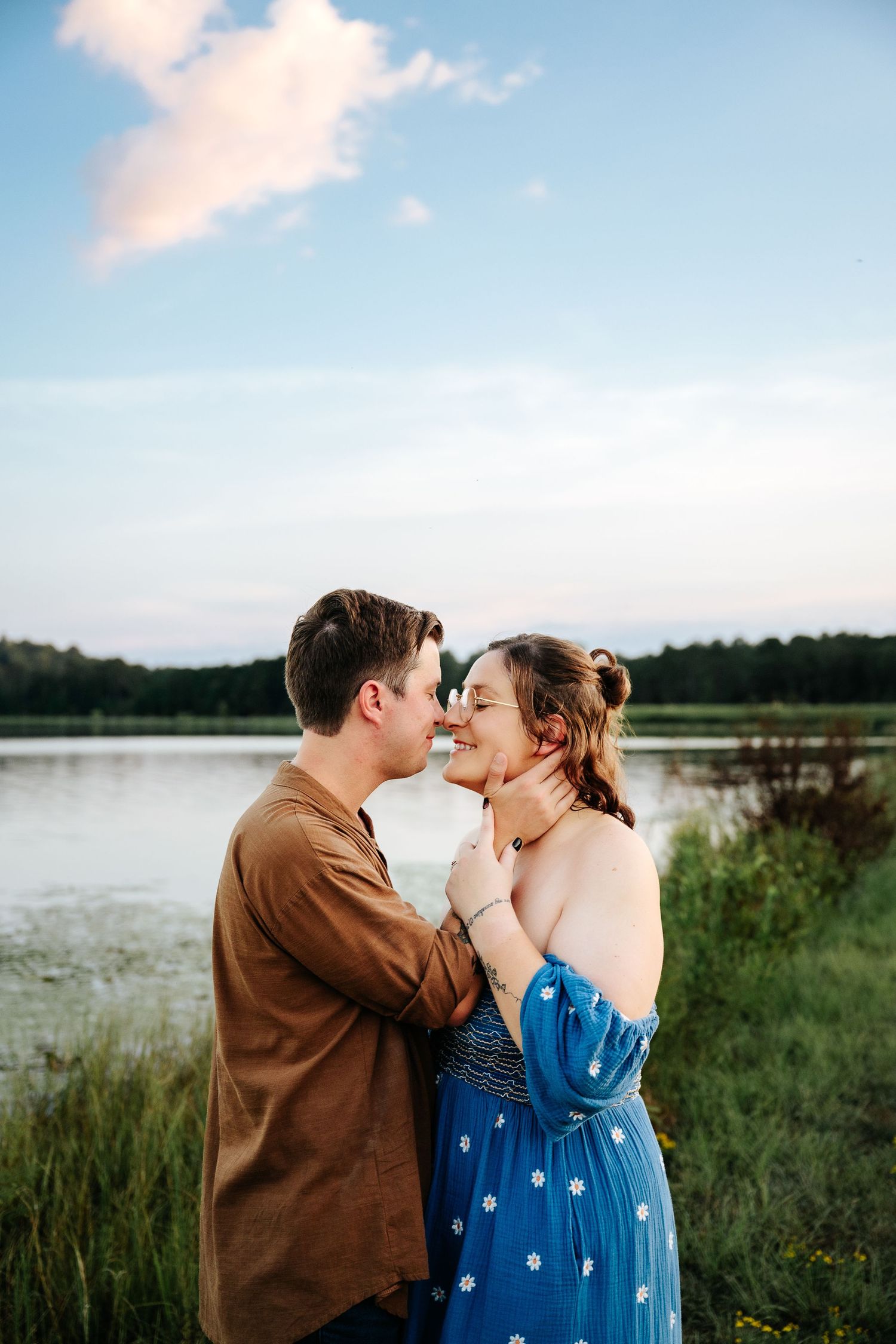 Couple sharing a romantic moment near a lake at sunset, woman in blue floral dress, man in brown shirt holding her face.