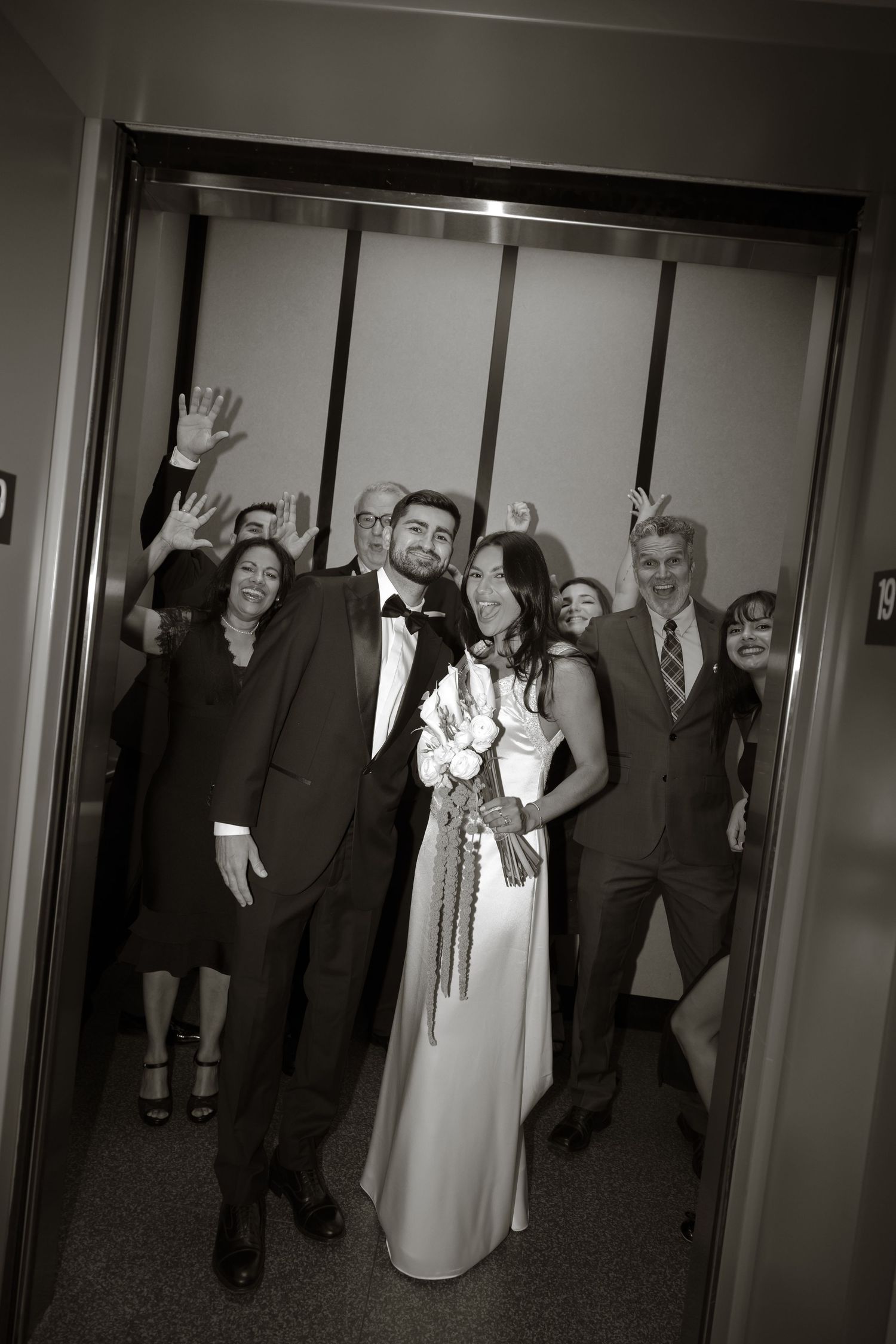 Black and white photo of a bride and groom posing in an elevator with joyful guests waving behind them at their wedding.