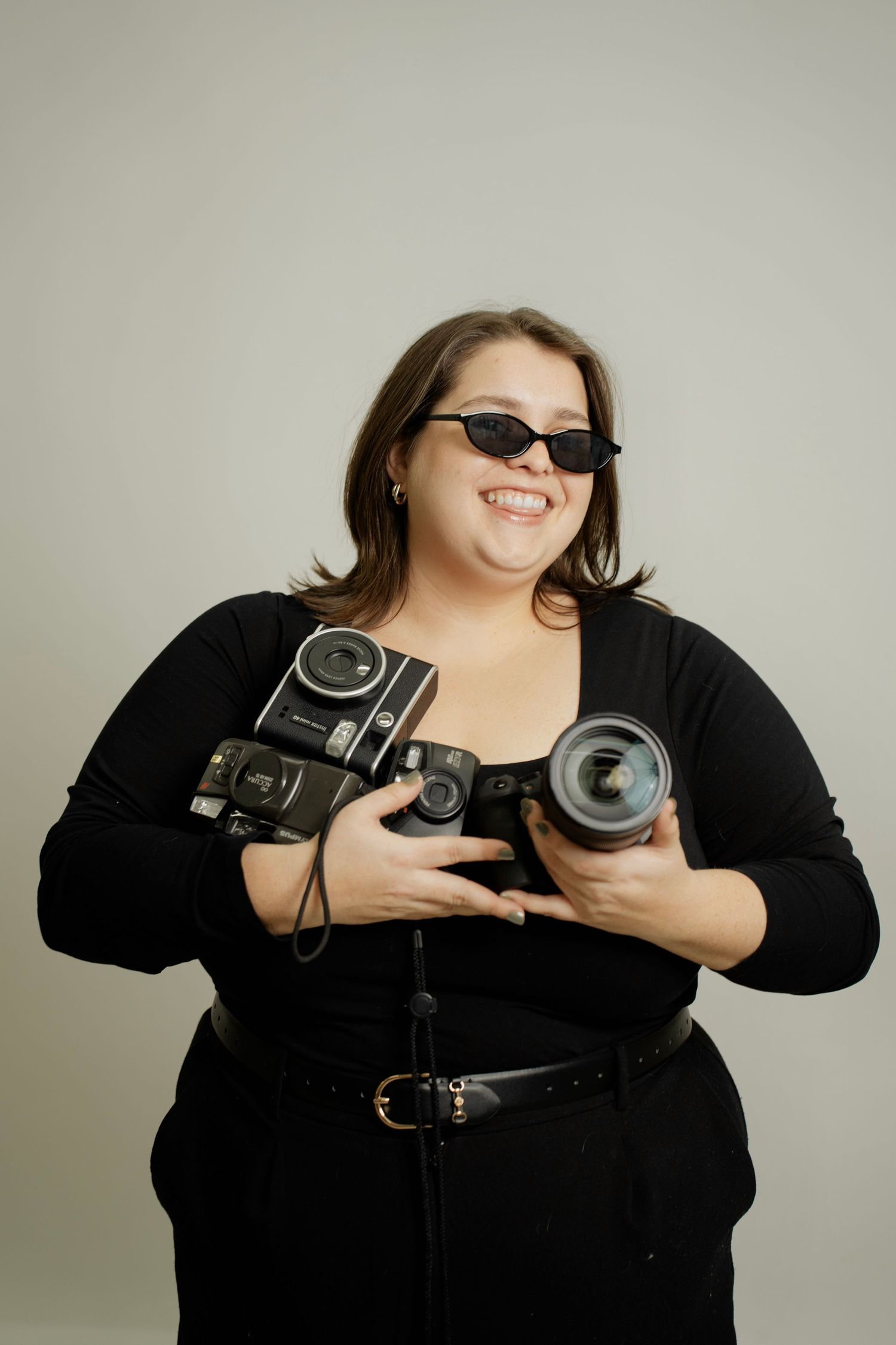 A photographer in black clothing holds various vintage cameras while posing against a light gray background.