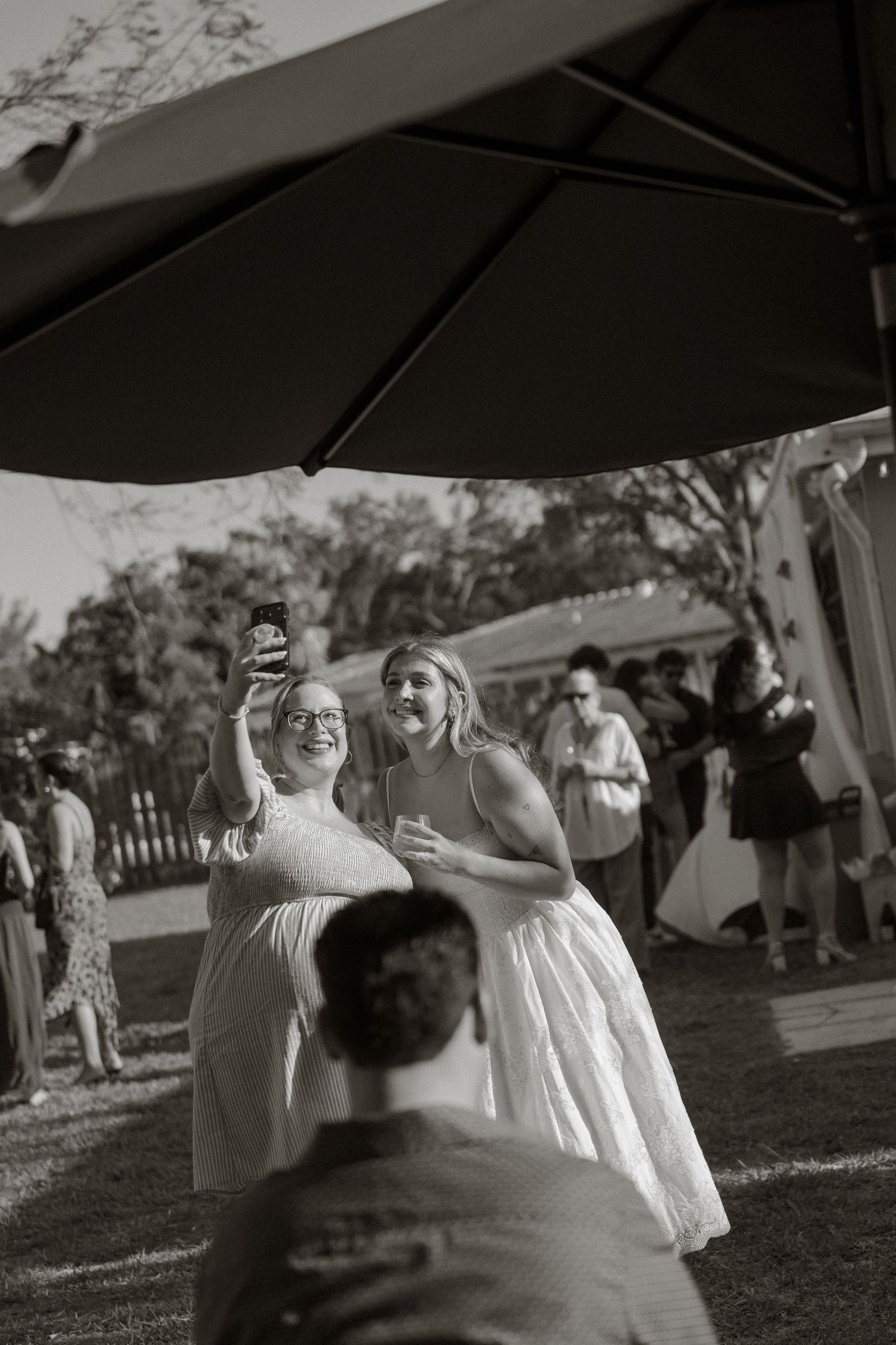 Two women in white dresses laugh while taking a selfie at an outdoor wedding reception in black and white photo.