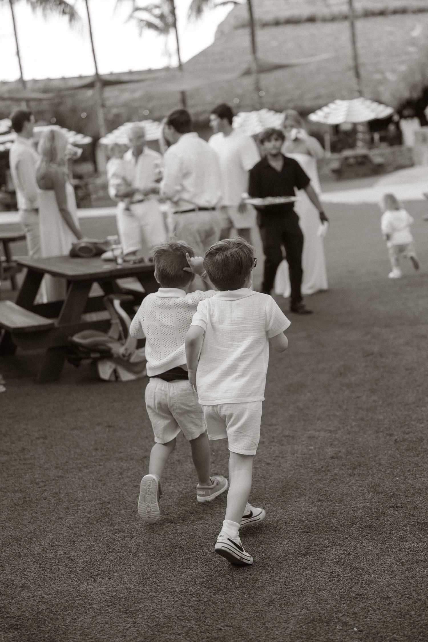 Two young boys in white outfits run together at an outdoor gathering with adults and picnic tables.
