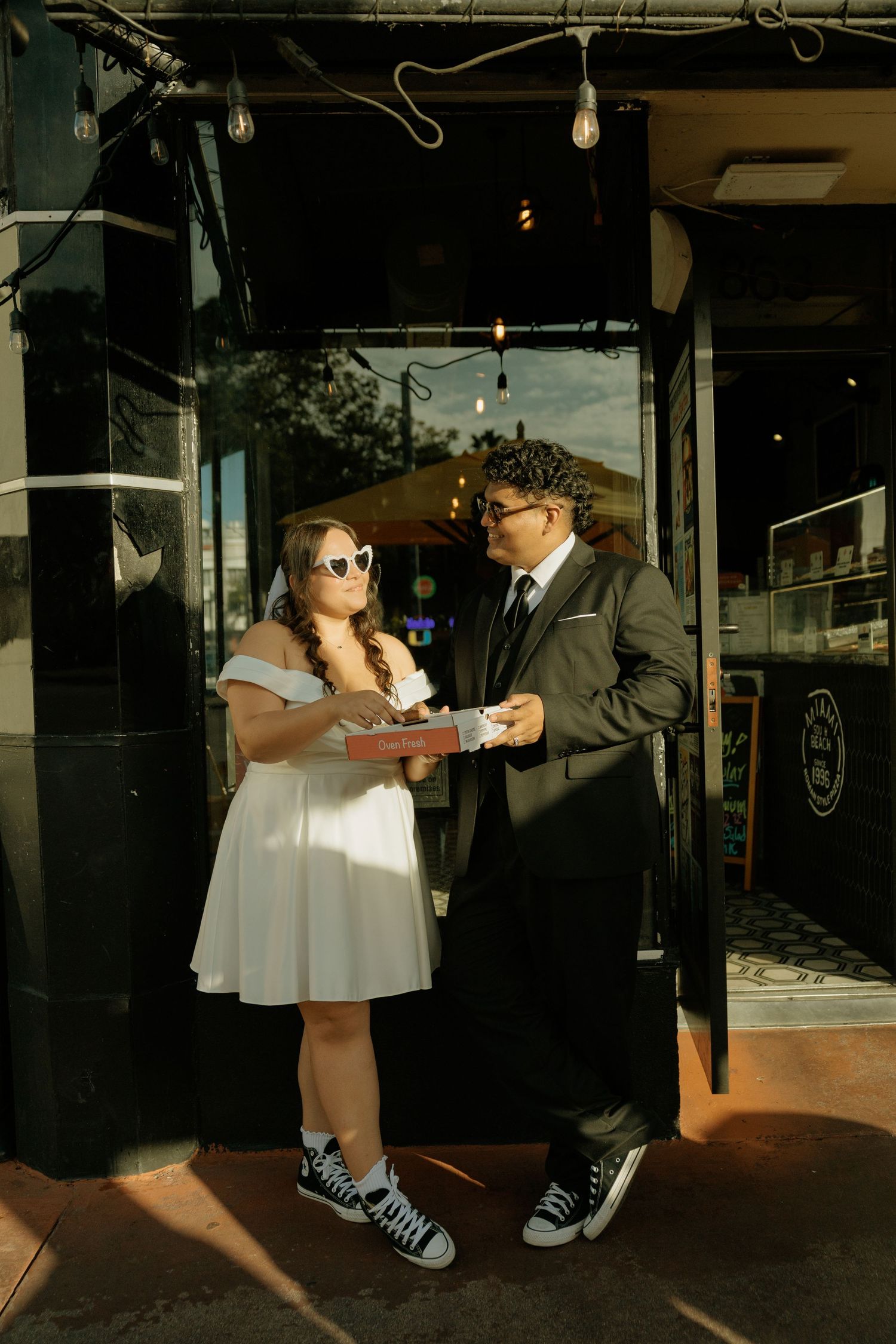 Couple in matching Converse sneakers sharing donuts outside a shop, woman in white dress and man in gray suit.