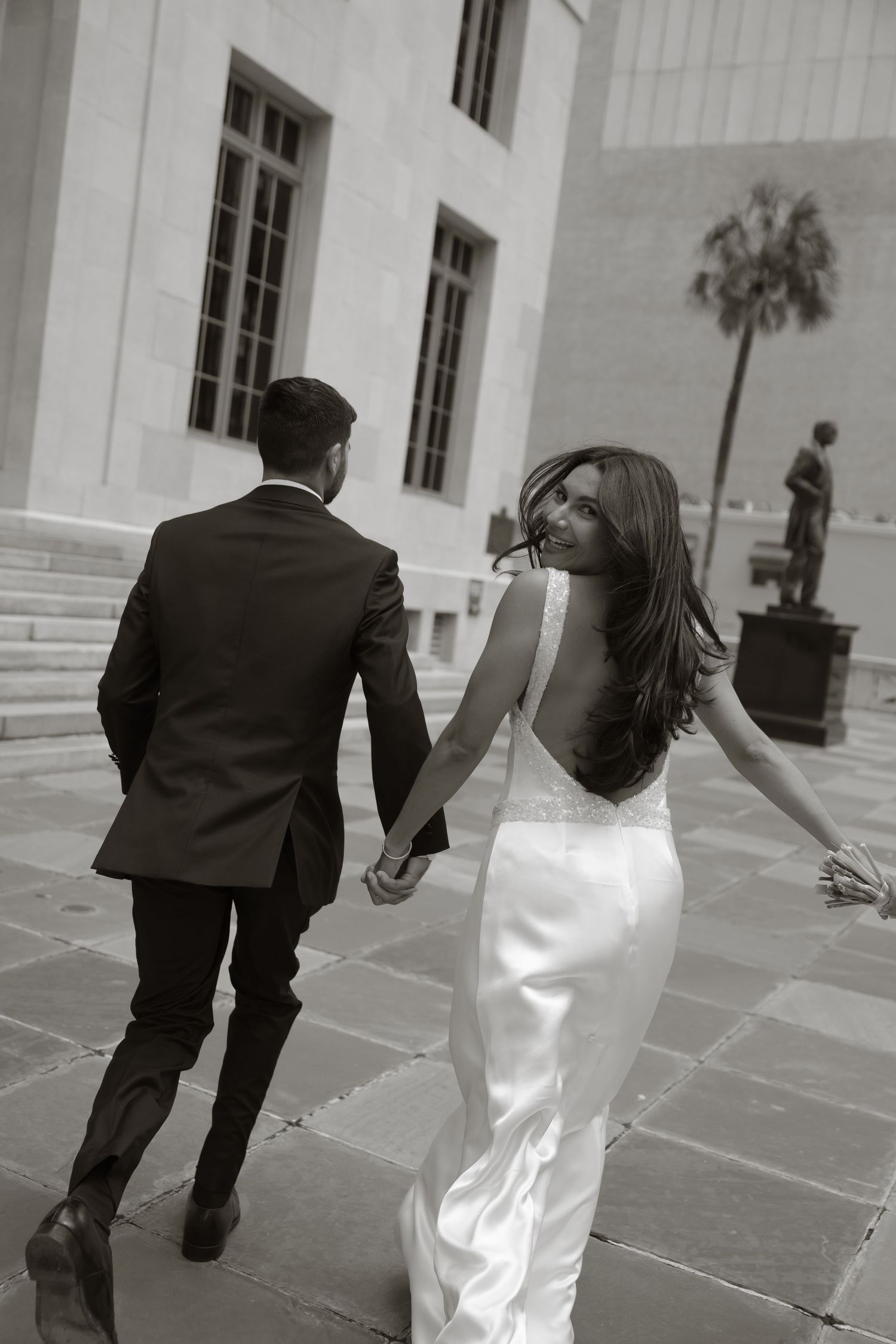 Black and white photo of a bride in a backless gown and groom in a suit holding hands walking toward a grand building.