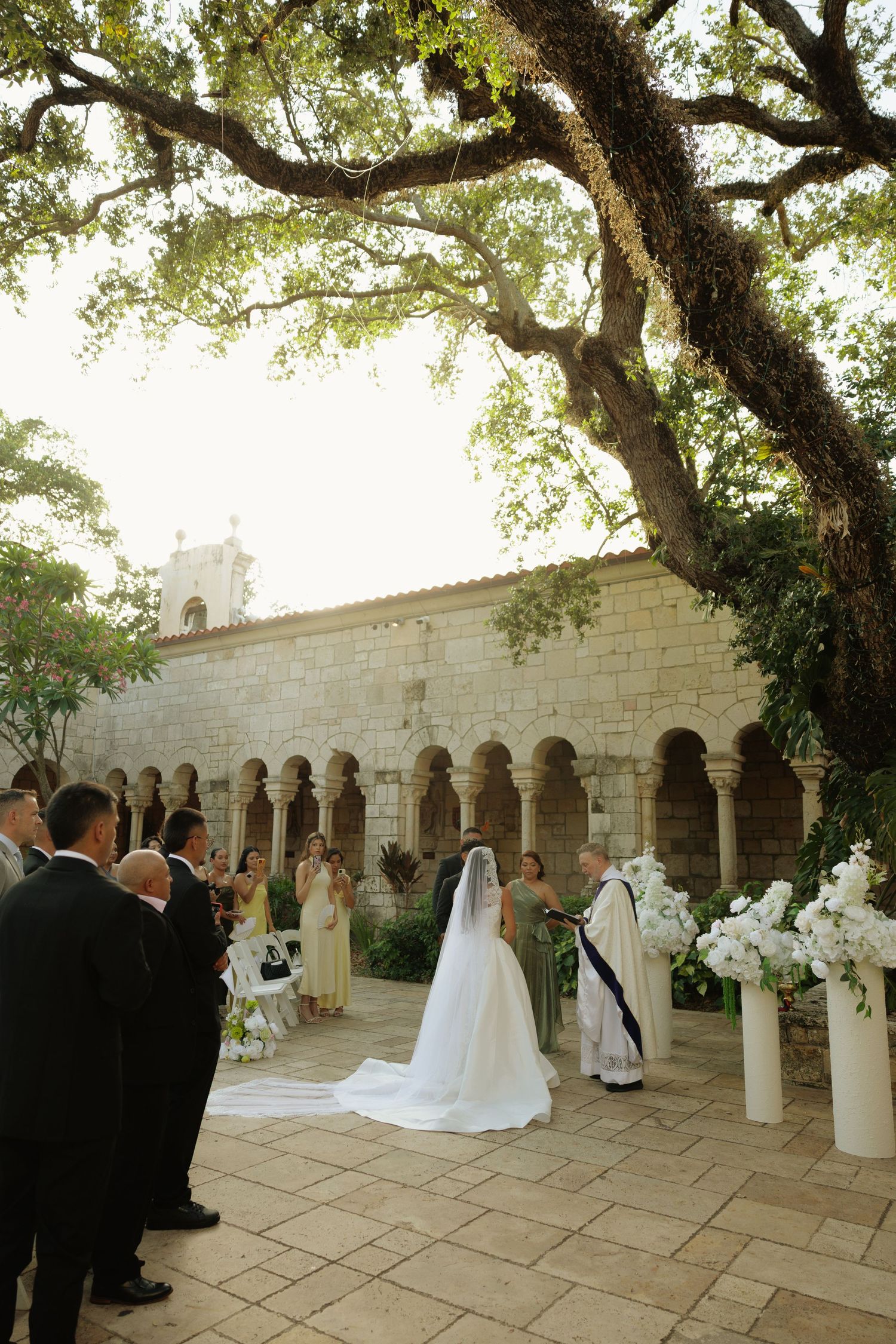 Outdoor wedding ceremony at historic stone chapel with bride in white gown and veil, white floral arrangements, and large oak tree.