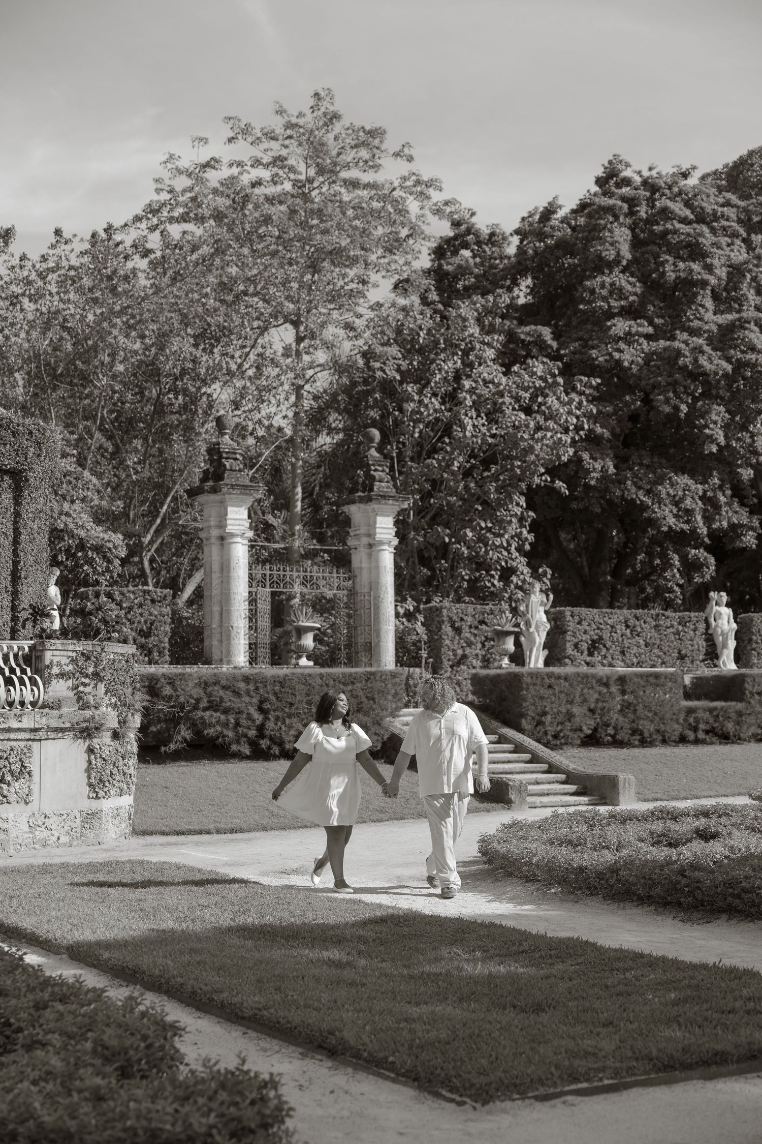Couple in white attire dancing in a formal garden with sculpted hedges and classical statues in black and white.
