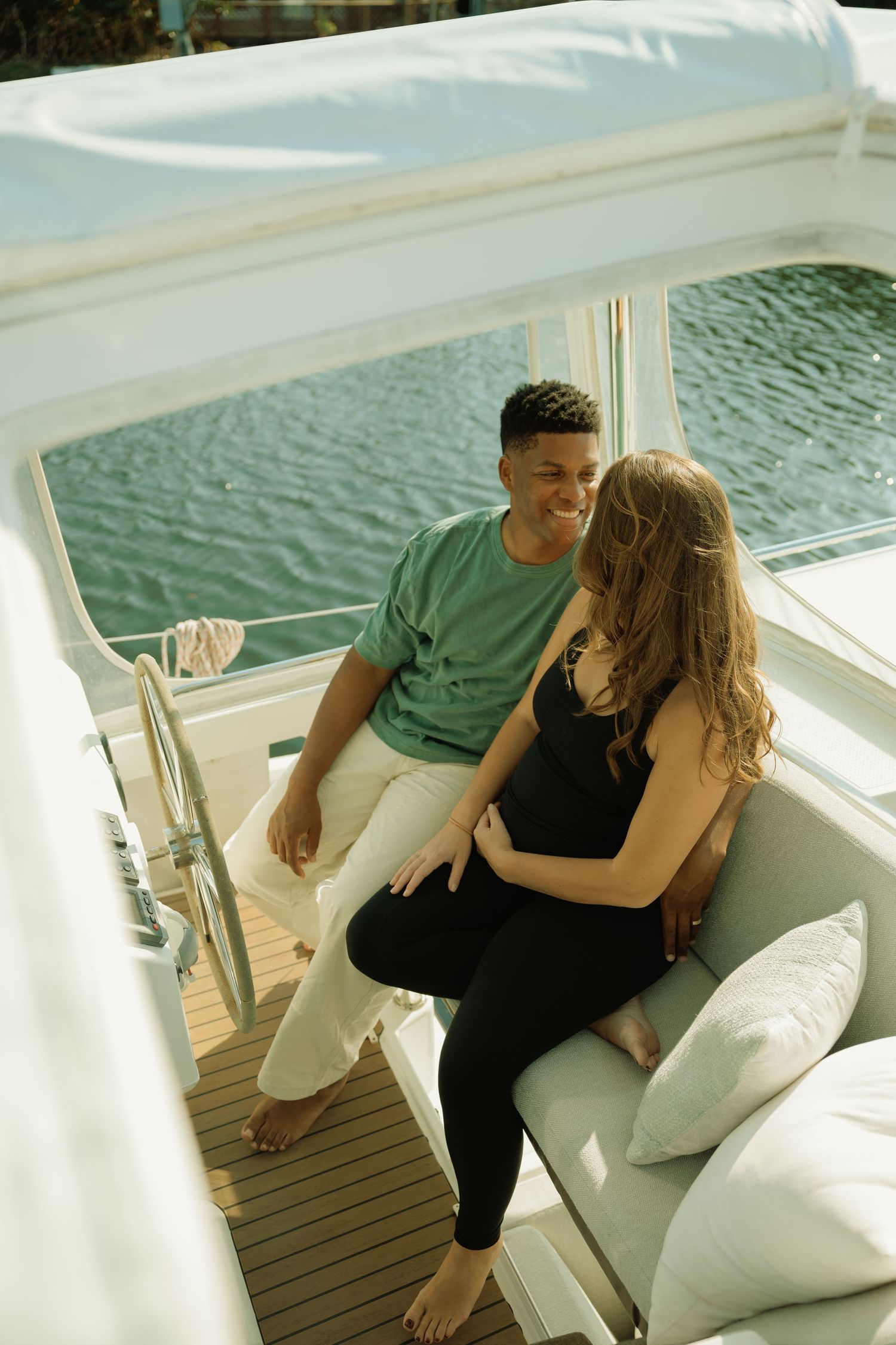A smiling couple sits together on a yacht deck, with the man in a green shirt and woman in black outfit.