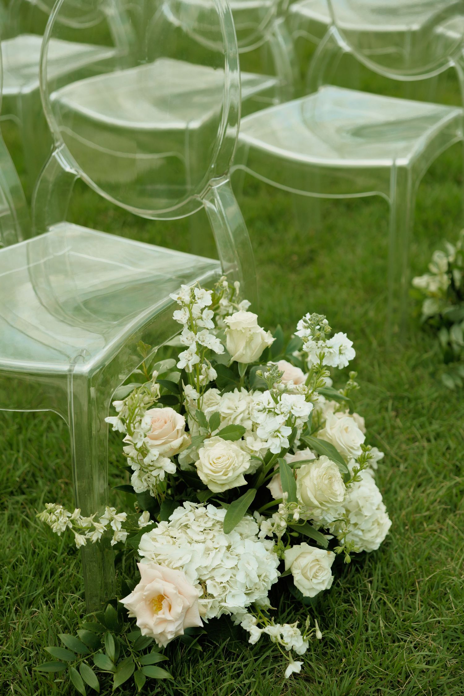 White and blush floral arrangement with roses and hydrangeas on green grass beside clear ghost chairs at outdoor wedding.