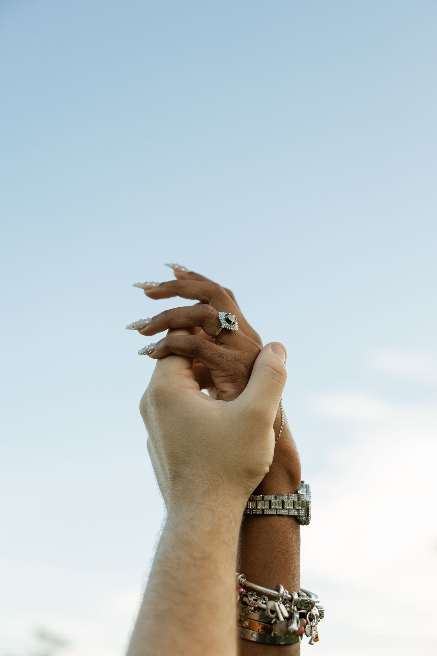 Hands adorned with multiple silver bracelets reaching up towards a bright blue sky.