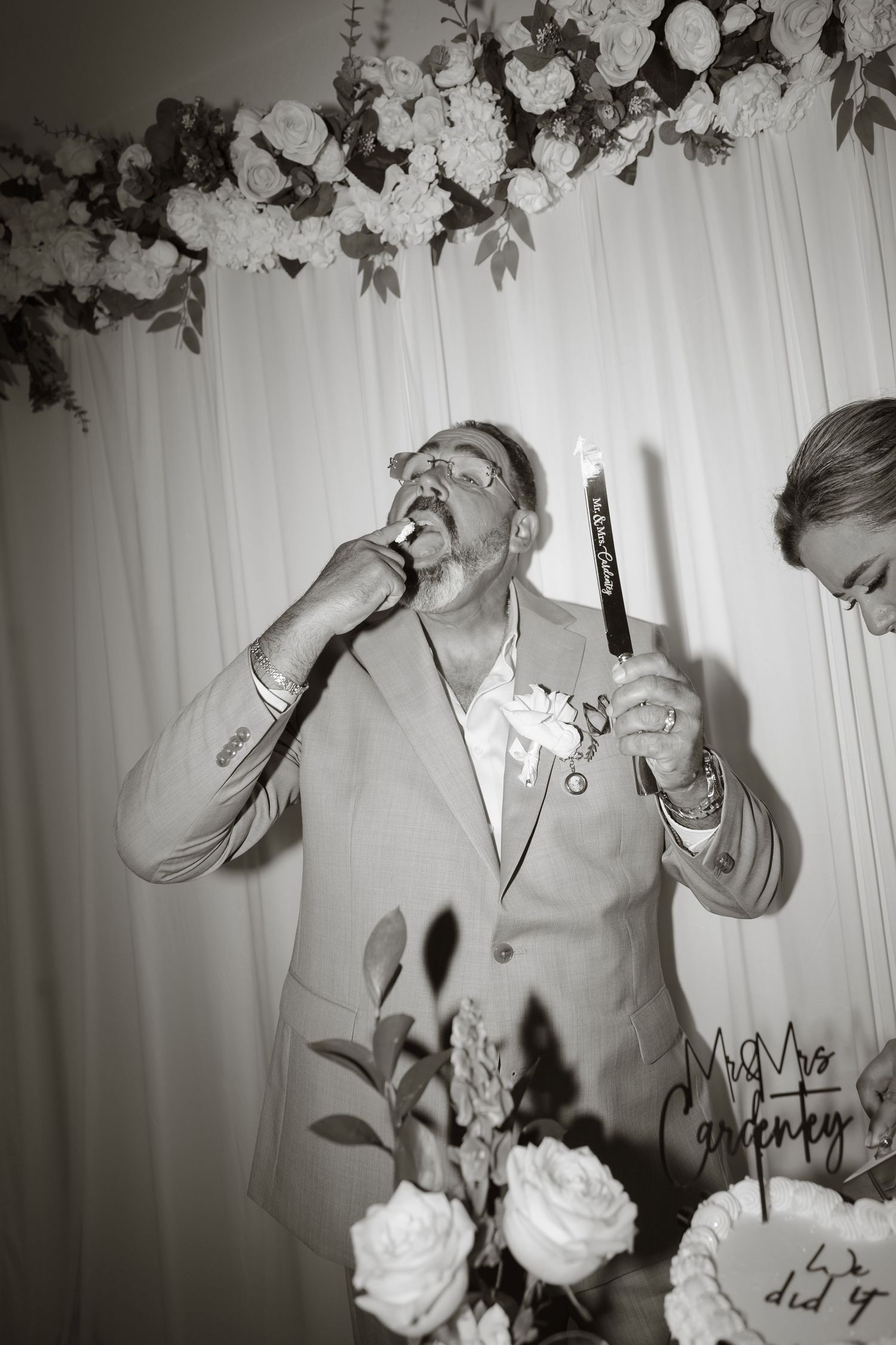 Groom in suit eating wedding cake at reception, floral backdrop with white roses, Mr & Mrs cake topper visible.