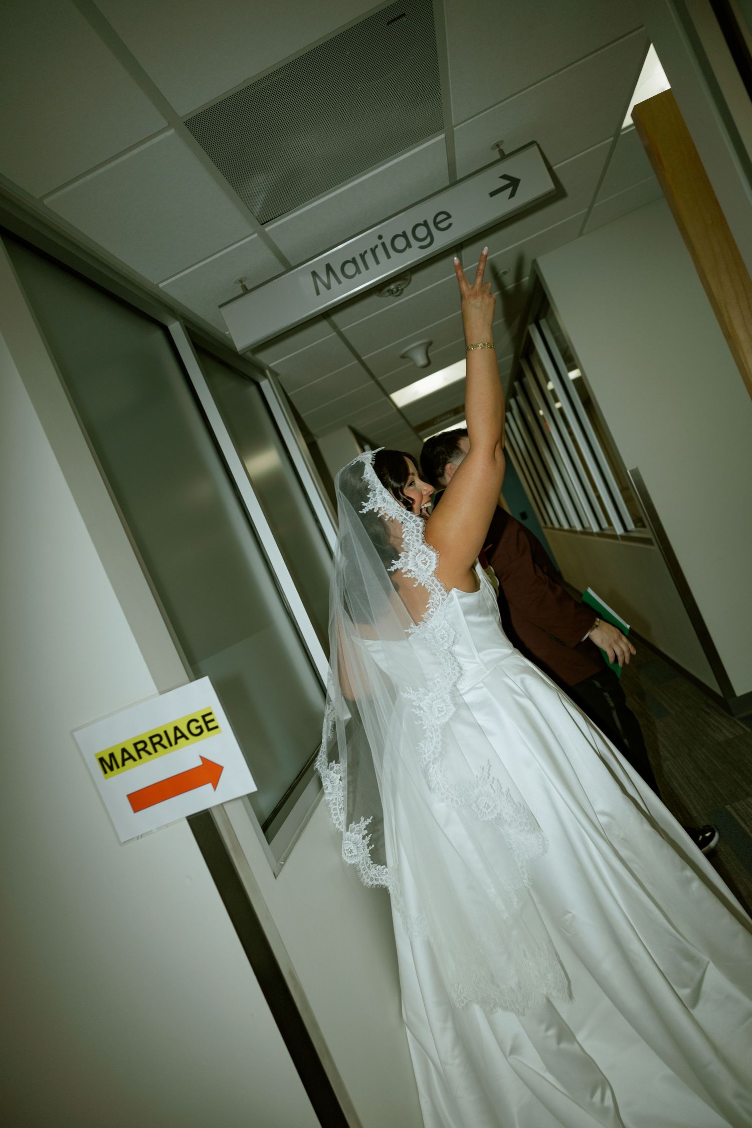 Bride in white gown and veil pointing at a Marriage sign in a hallway, with groom beside her.
