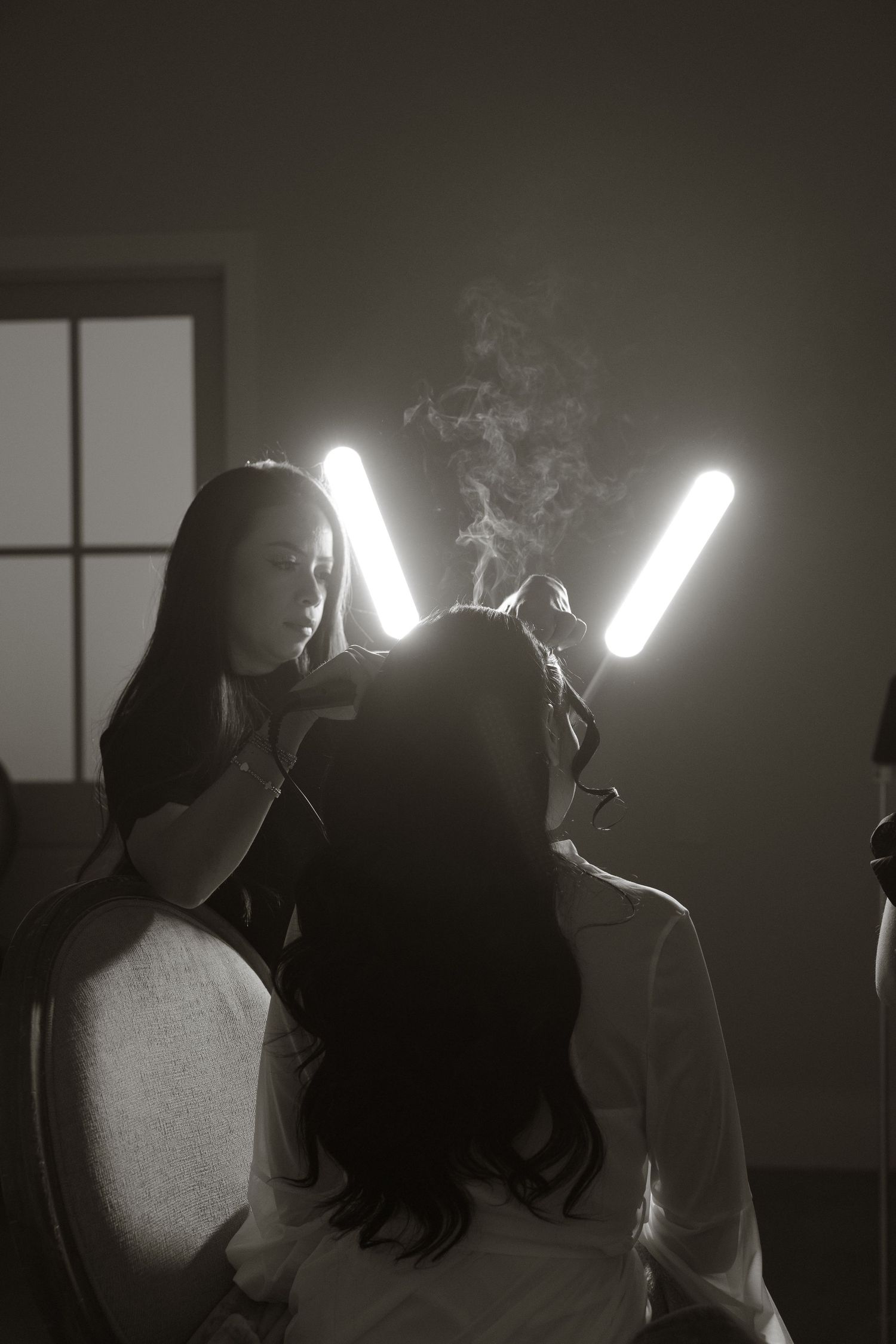 Black and white photo of a hairstylist using hot tools on a client, with steam rising under bright tube lights.
