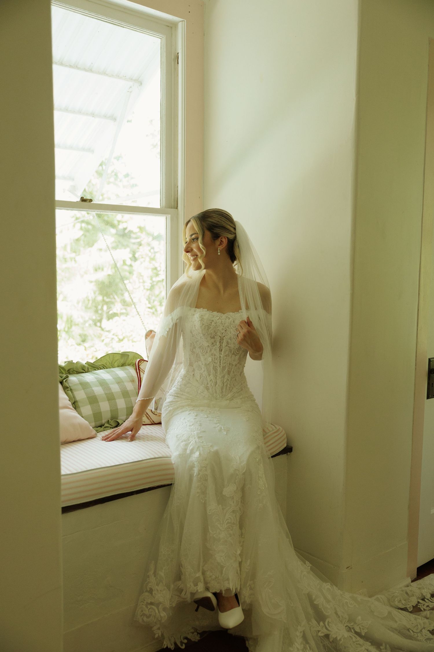 Bride in lace wedding gown and veil sits on window seat, gazing outside at greenery in soft natural light.