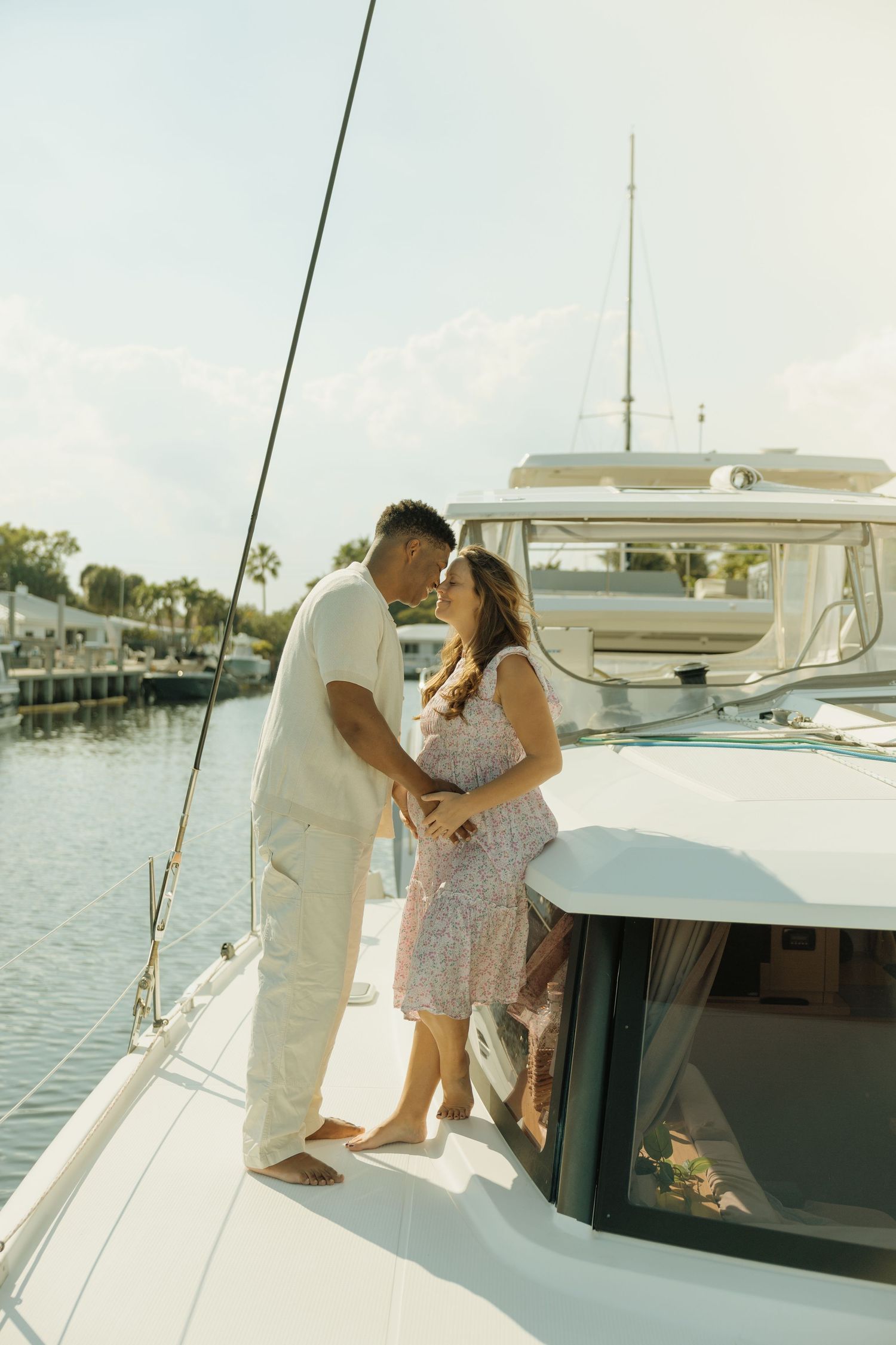 A romantic couple embraces on the deck of a white sailboat in a sunny marina, with calm waters and trees in the background.