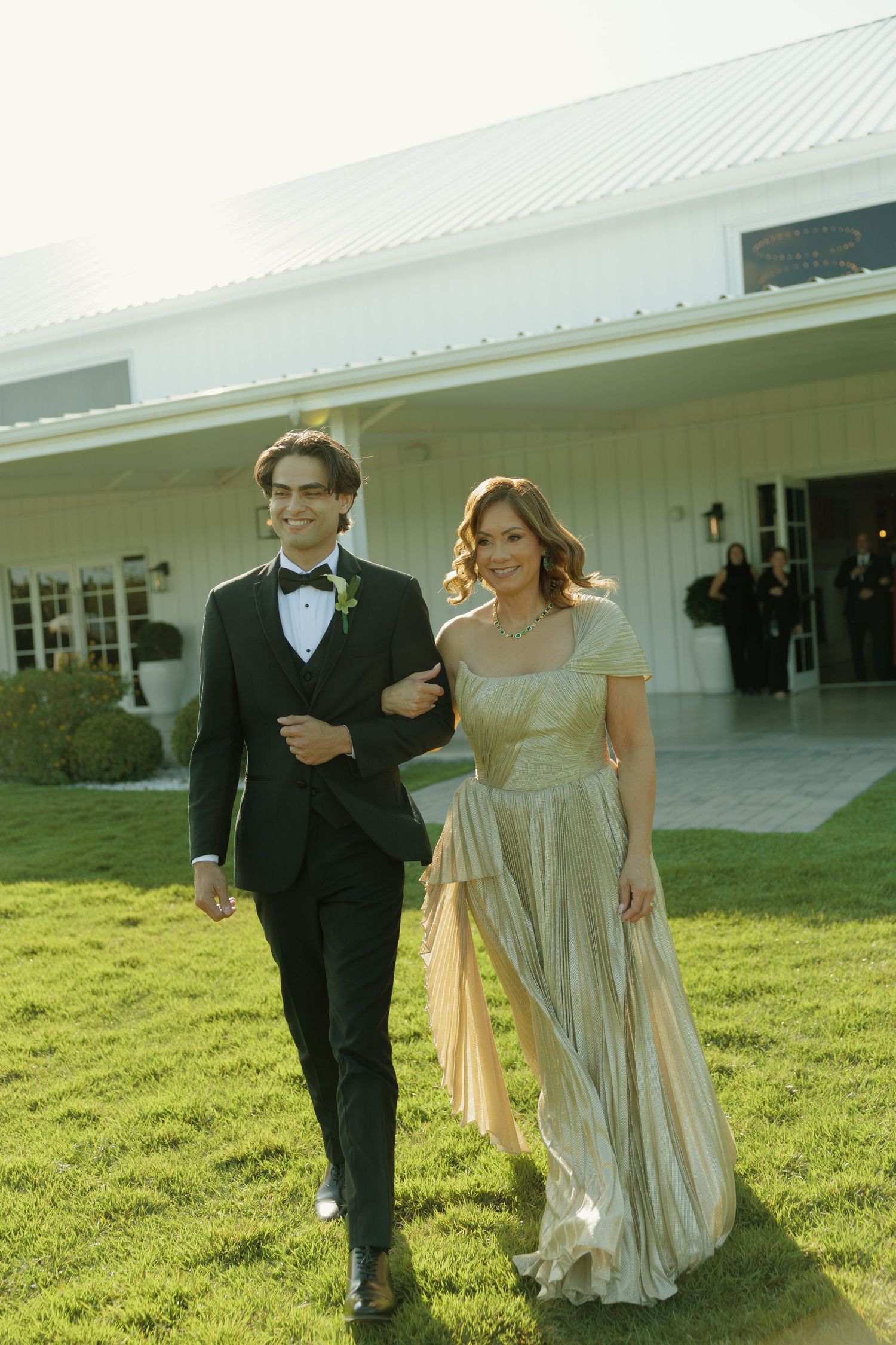 Elegant couple walking outdoors, man in black tuxedo and woman in gold gown, at a formal event near white building.
