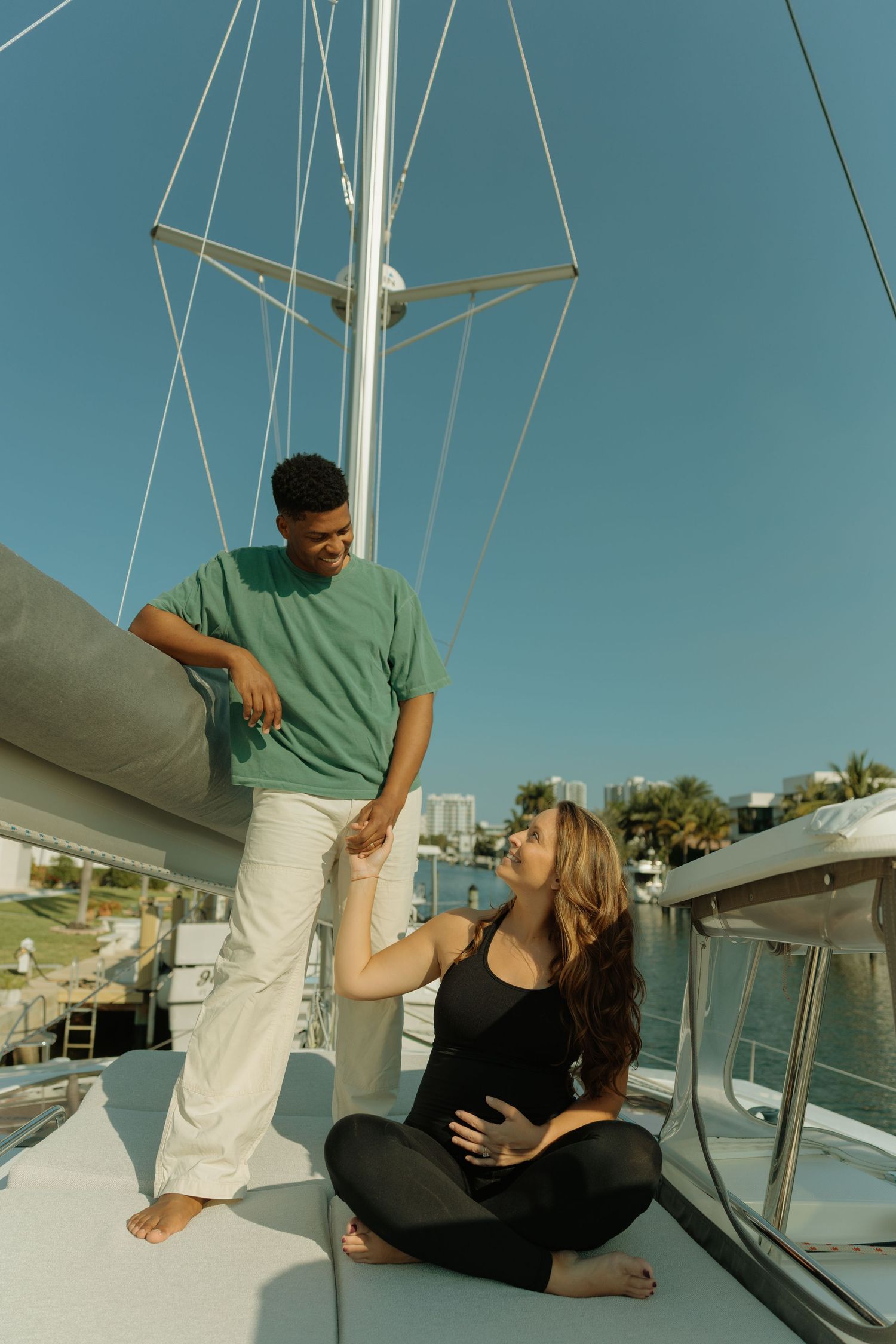 A couple relaxes on a sailboat deck, man in green shirt holds woman's hand as she sits cross-legged in black outfit.
