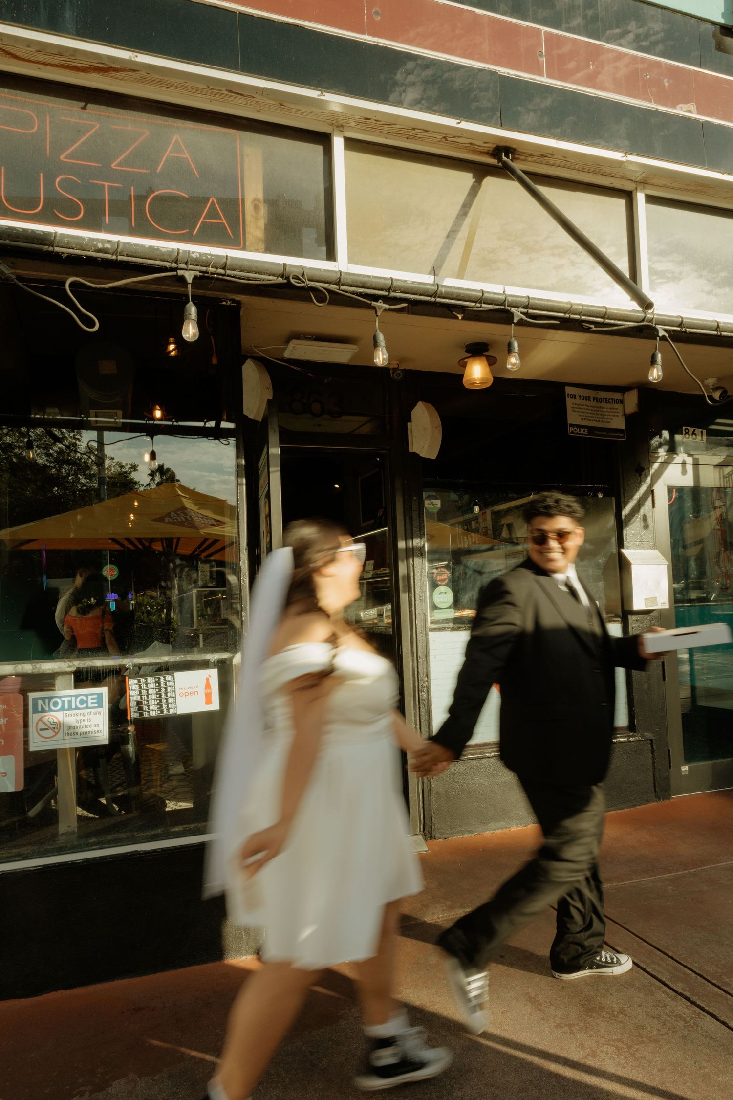 Bride and groom walking hand in hand past Pizza Rustica restaurant in motion-blurred wedding photo.