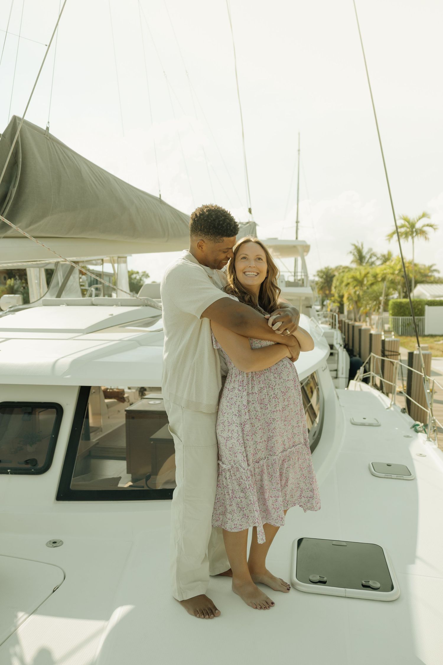 A couple embraces on a white sailboat deck, with the man kissing the woman's cheek near tropical palm trees.