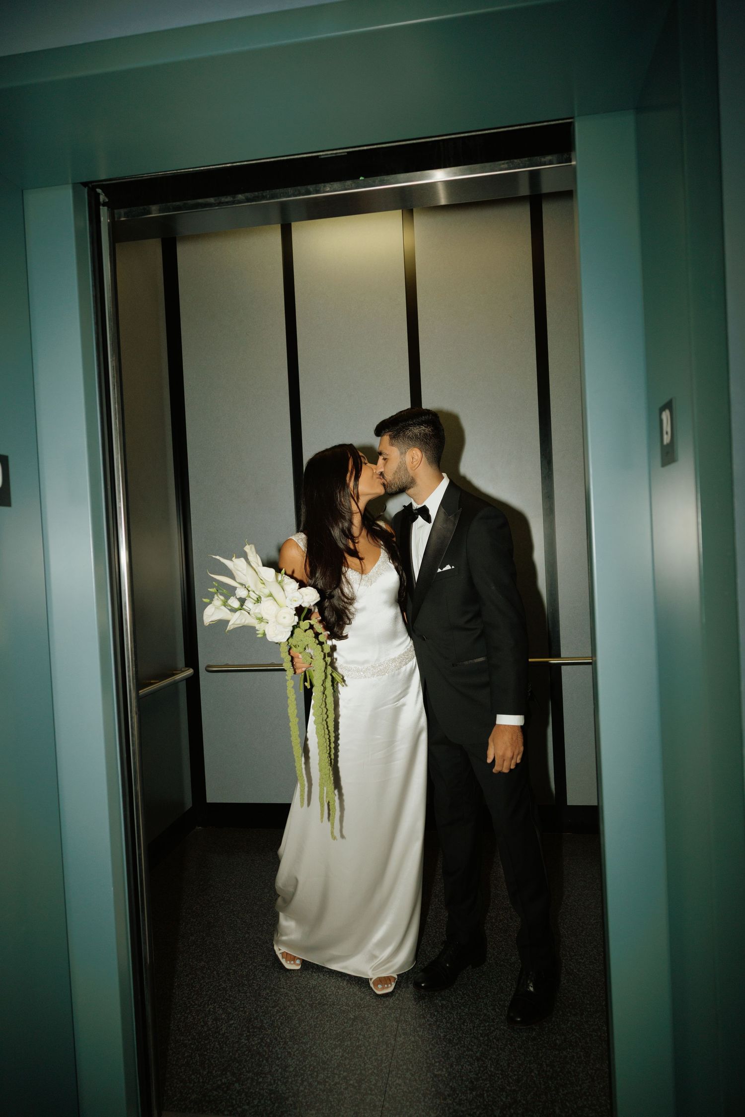 Bride and groom sharing a kiss in an elevator, bride holding white floral bouquet, groom in black tuxedo.