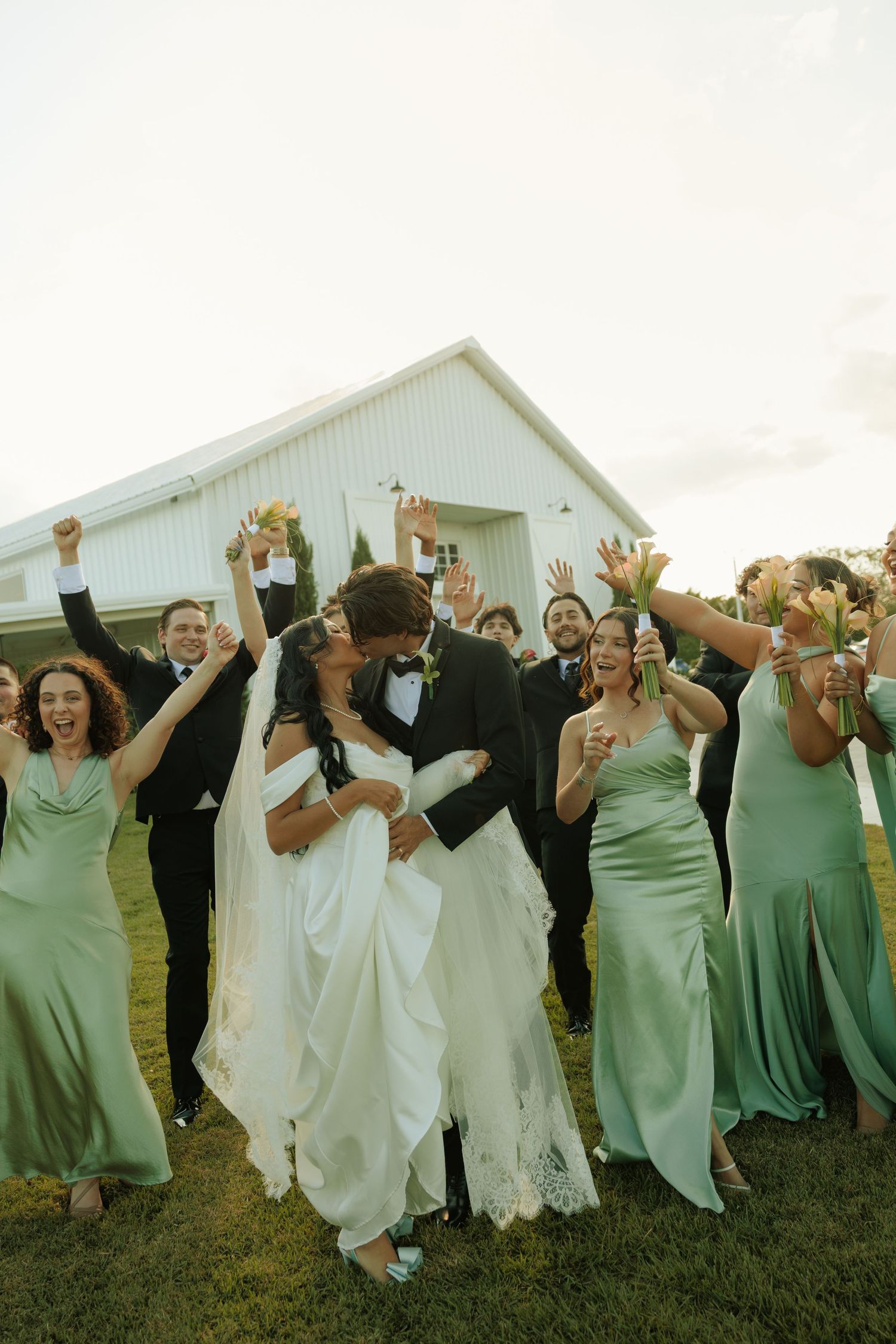 A joyful wedding party celebrates as the bride and groom share a kiss outdoors near a white barn.
