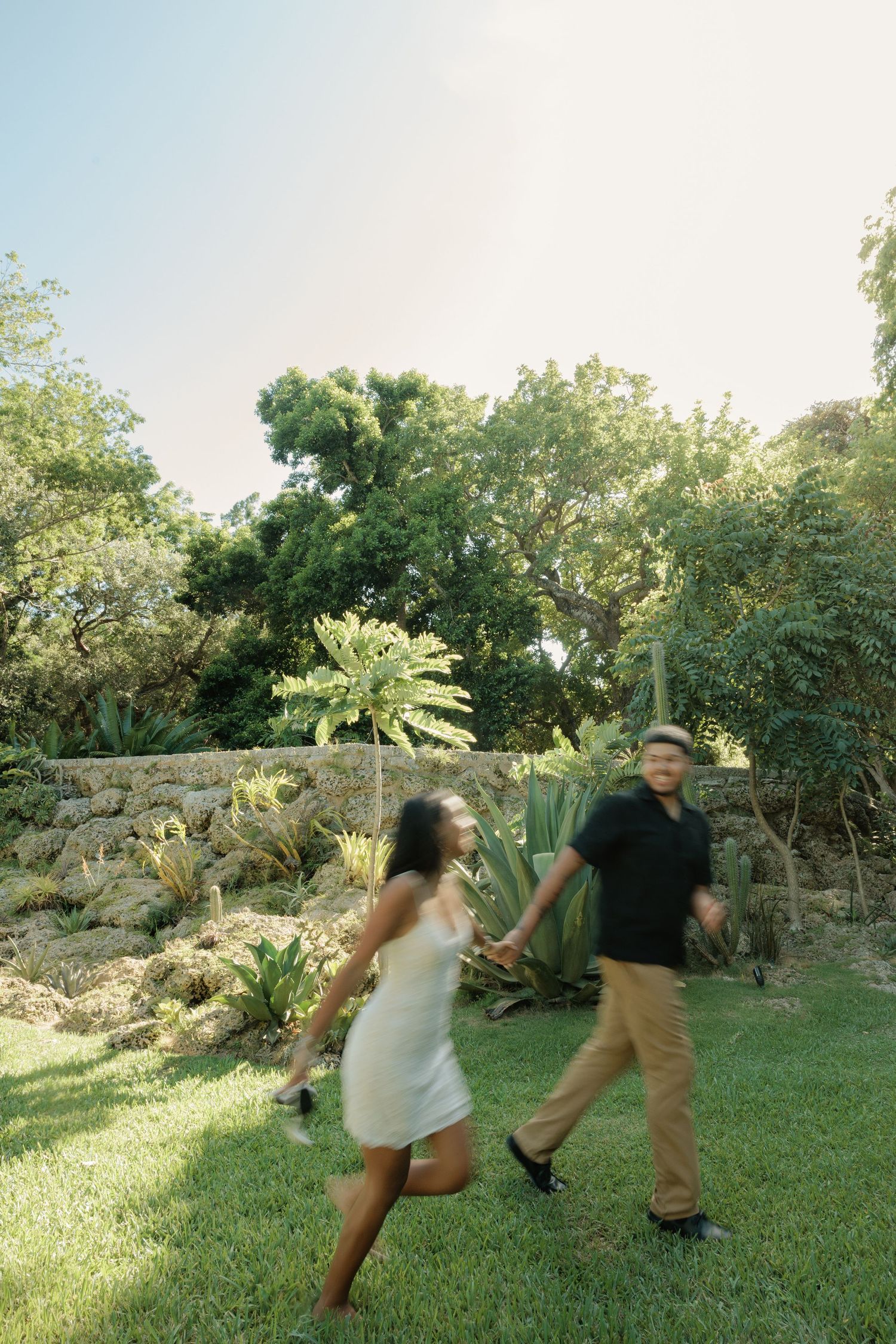 Two people holding hands and walking on grass in a lush garden setting with stone walls and tropical plants.