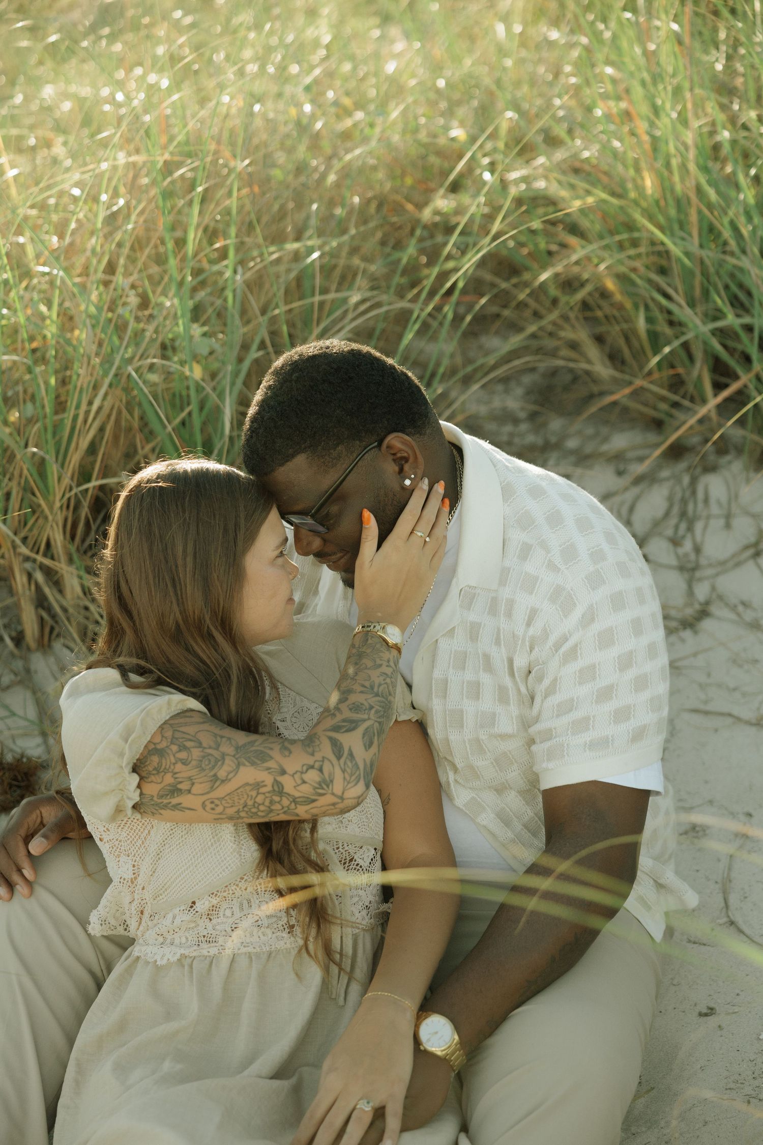 A romantic couple embraces while lying on sandy beach dunes during a tender moment at sunset.