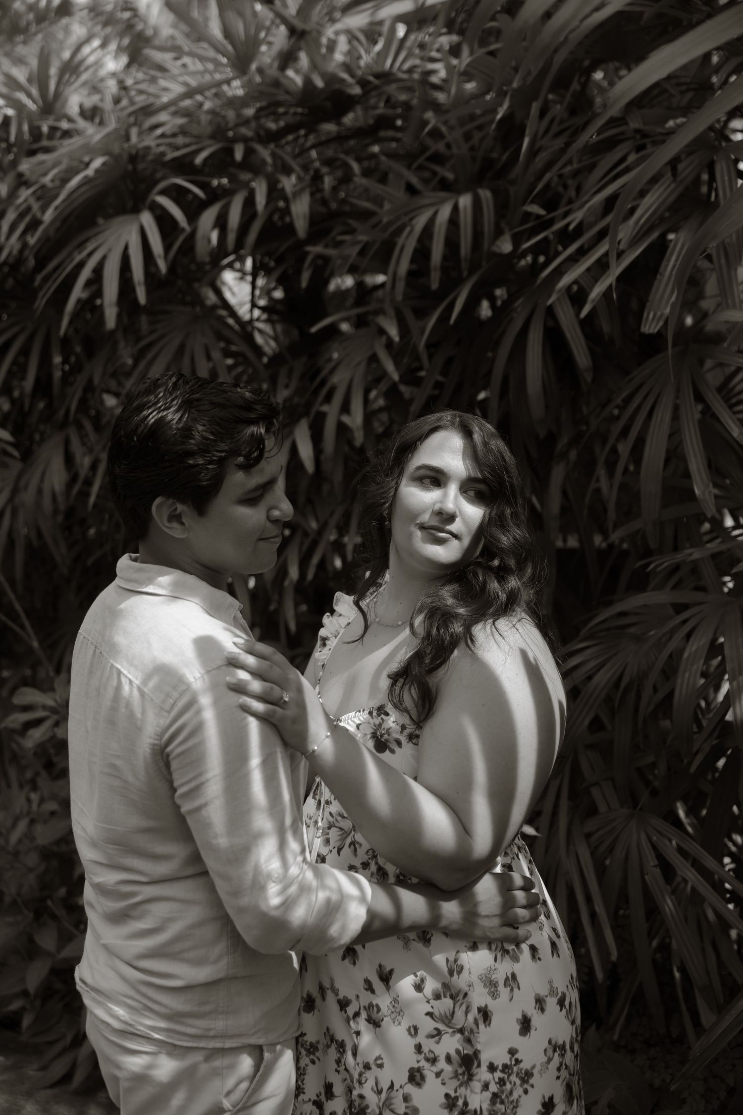 Black and white photo of a couple embracing romantically in front of lush tropical foliage.