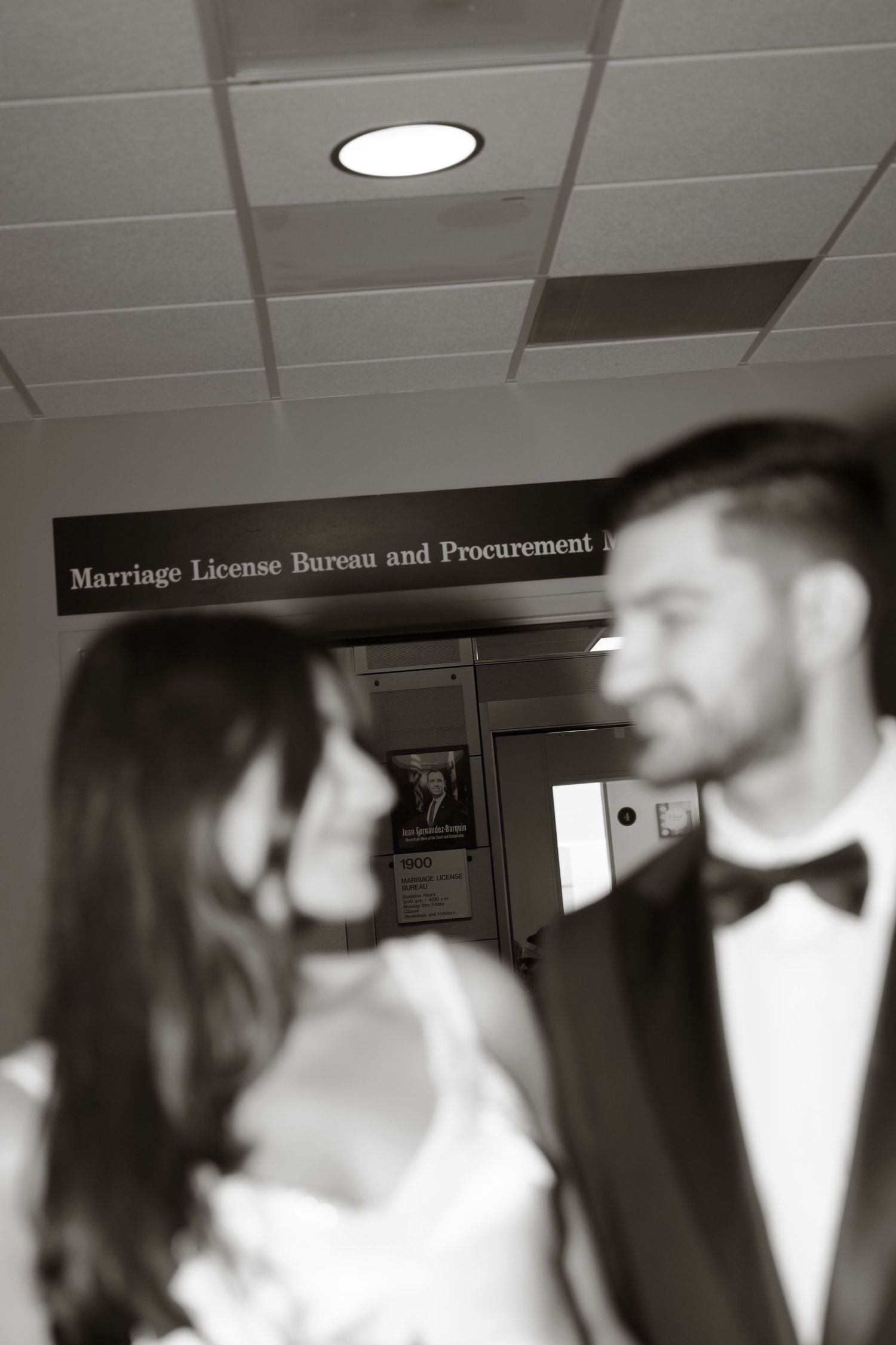 Black and white photo of a couple in wedding attire standing beneath a Marriage License Bureau sign.