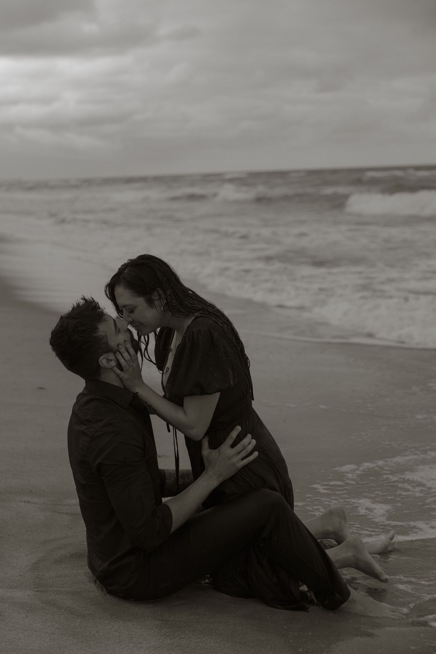 Black and white photo of a romantic couple kissing on a beach shore with ocean waves in the background.