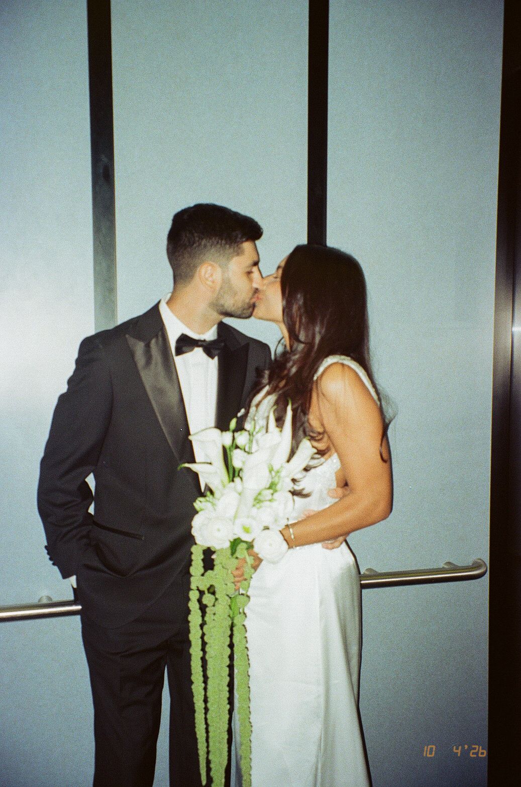 A couple shares a kiss on their wedding day, groom in black tuxedo, bride in white gown holding white floral bouquet.