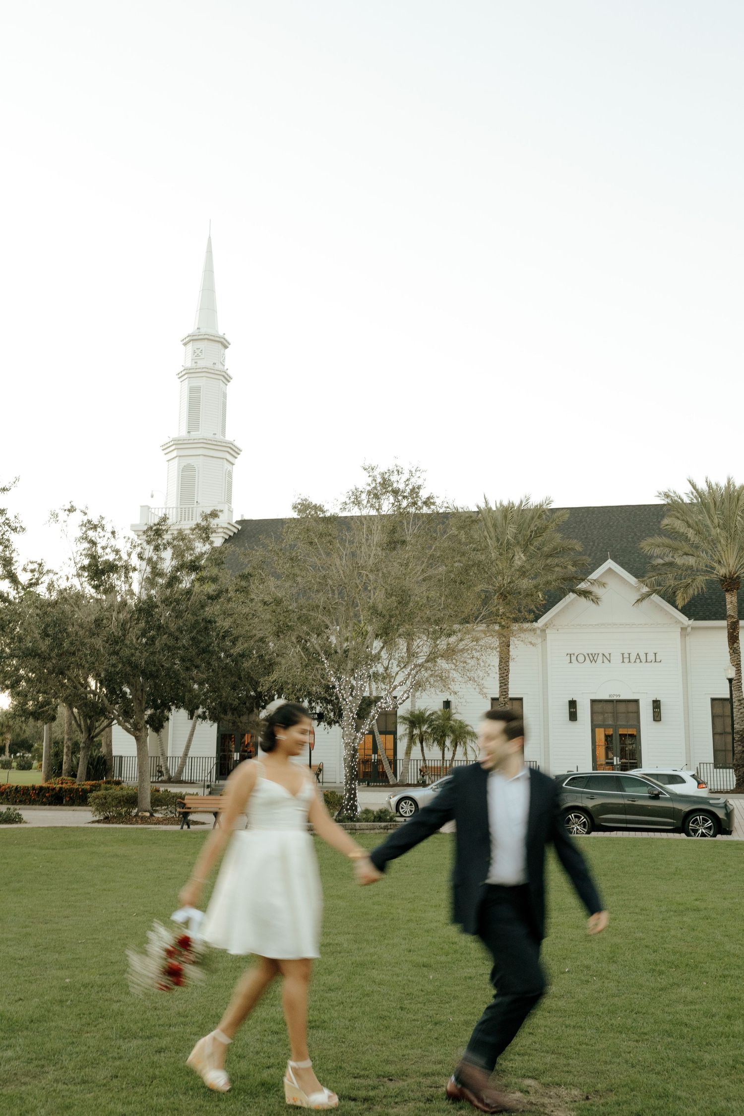 A couple joyfully dances and spins on a grassy lawn with a white town hall building and church steeples in the background.