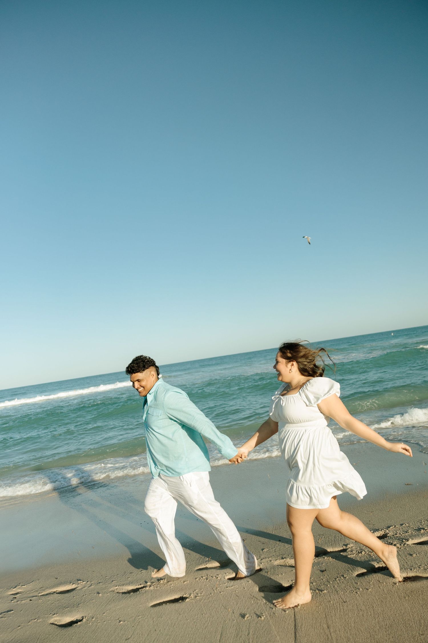 Two people in white outfits dance playfully along a sandy beach with turquoise ocean waves in the background.