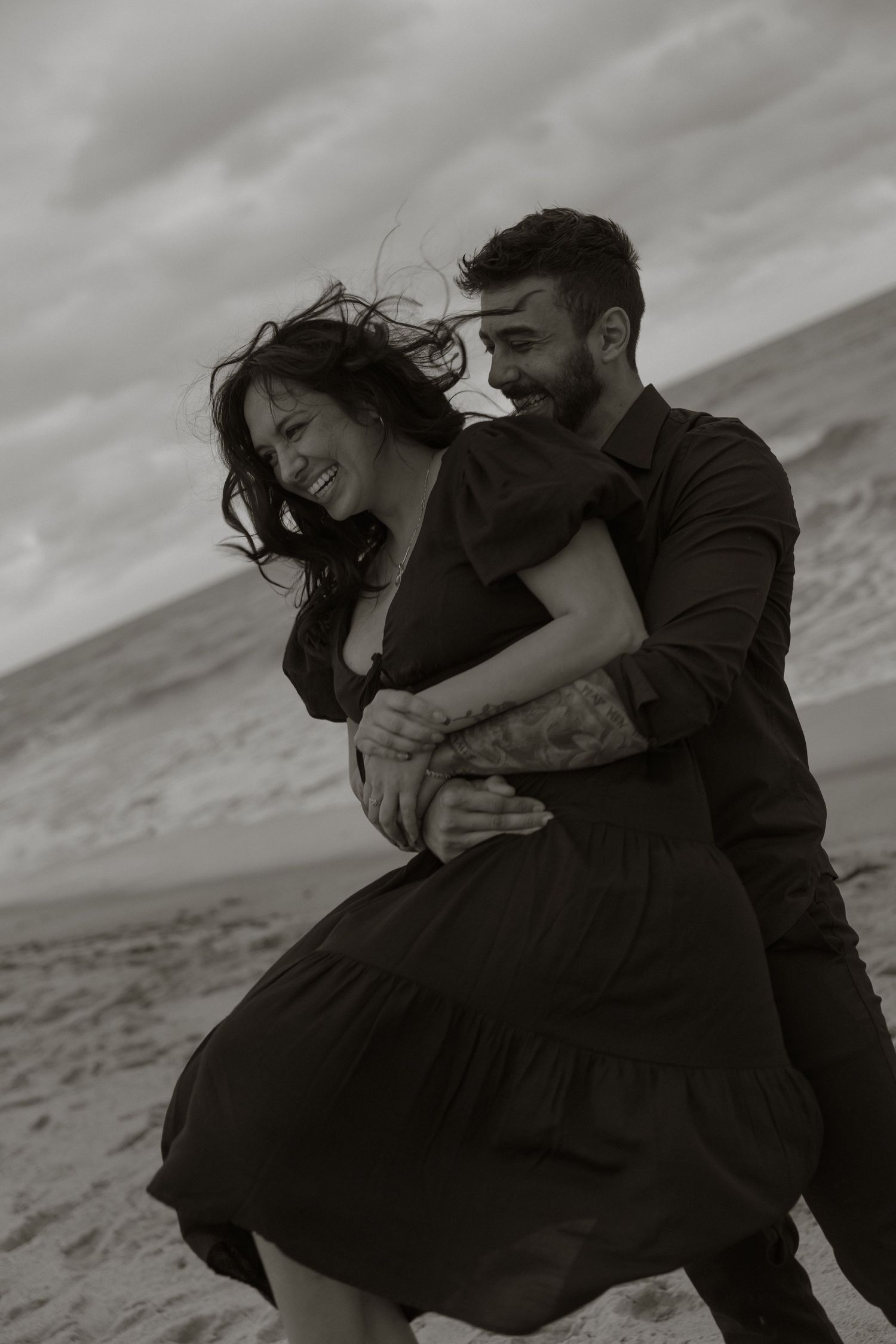 A romantic black and white photo of a couple embracing on a beach with wind-blown hair and dramatic cloudy sky.
