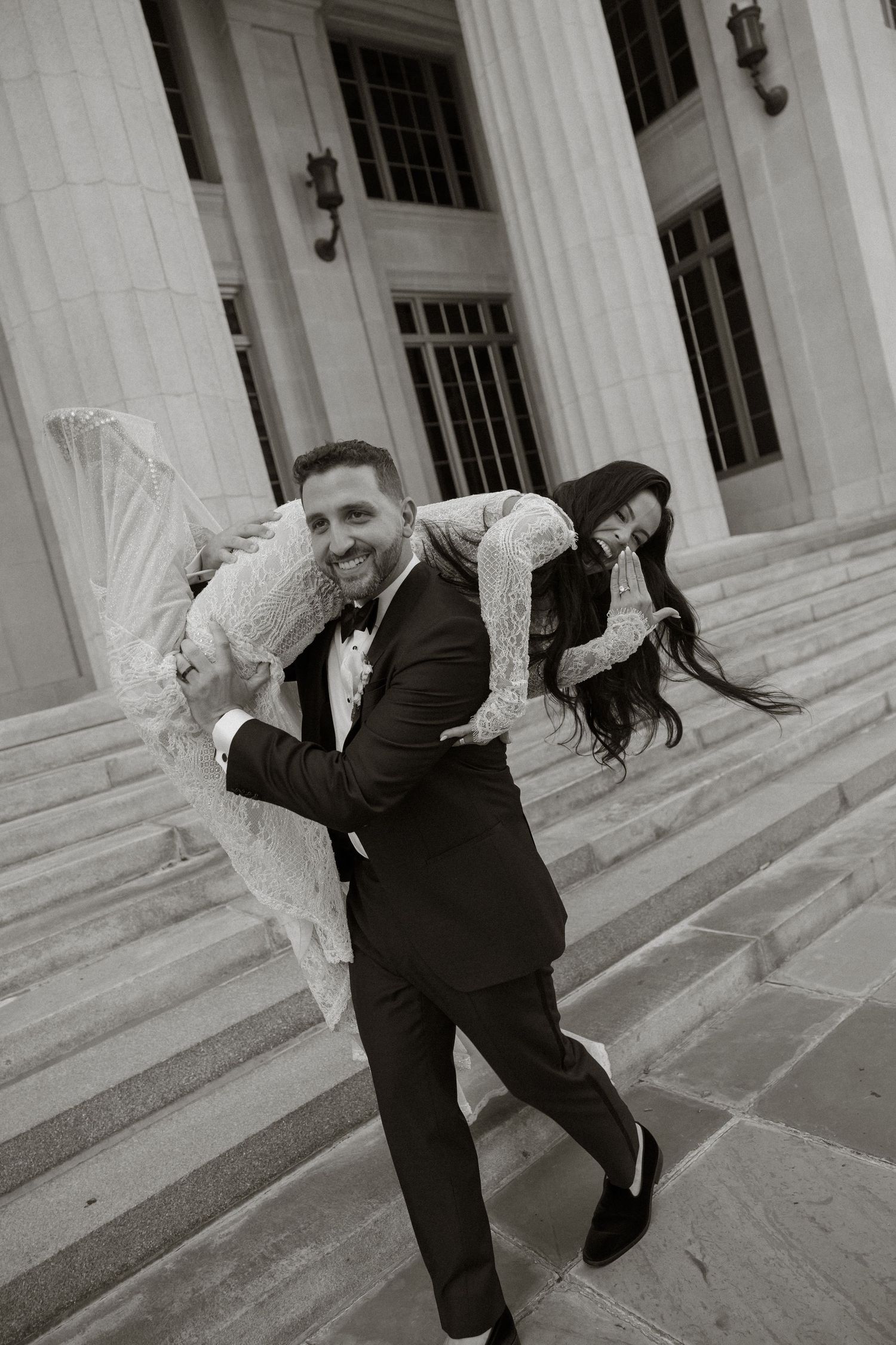 Groom in tuxedo carries bride in white lace dress over his shoulder on grand stone steps in black and white photo.