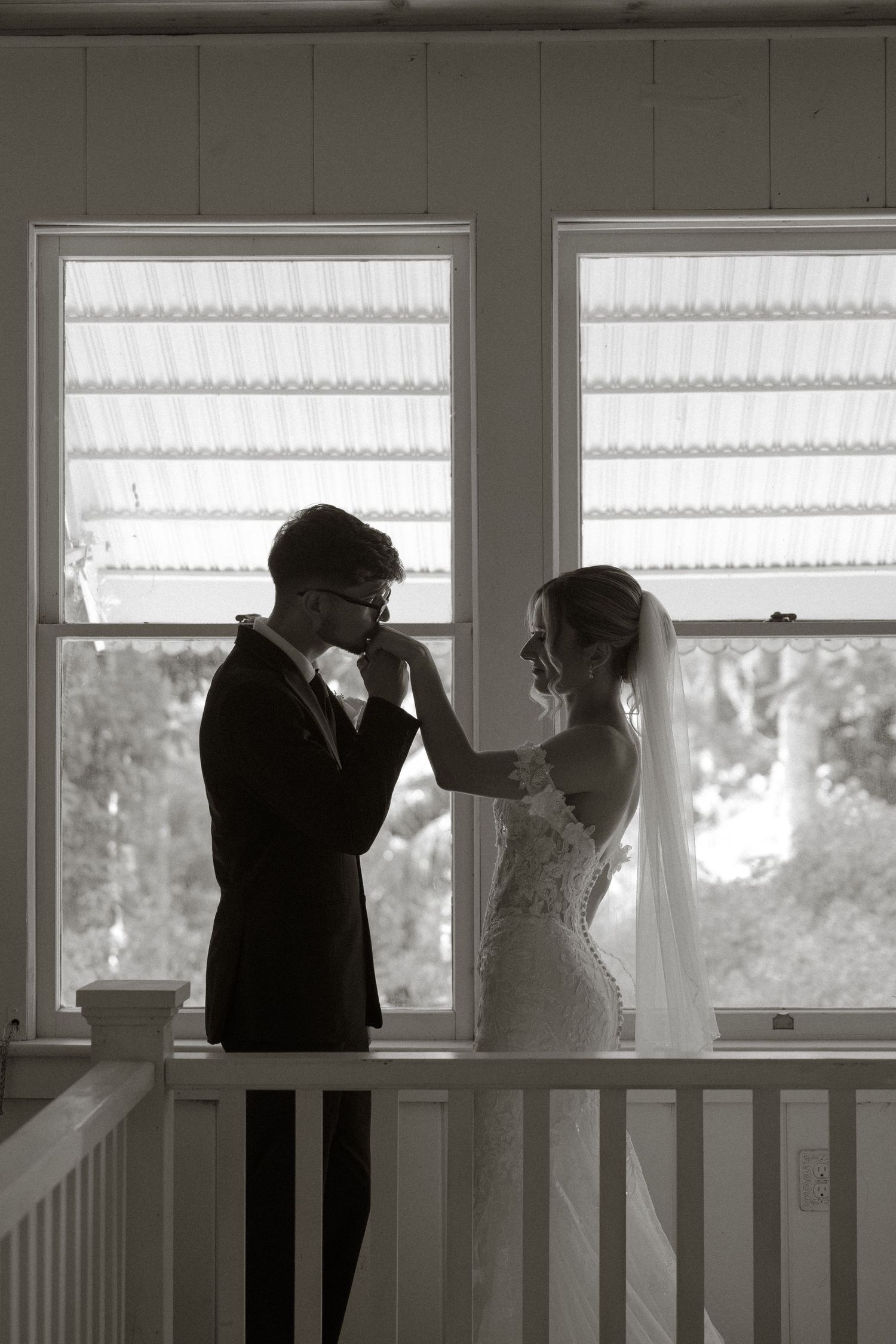 Black and white photo of groom kissing bride's hand by sunlit windows on wedding day.