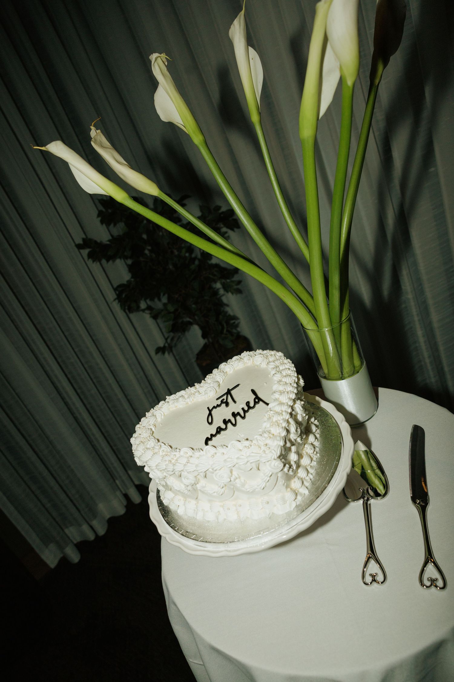 Heart-shaped white wedding cake with Just Married inscription beside calla lilies on a white table.