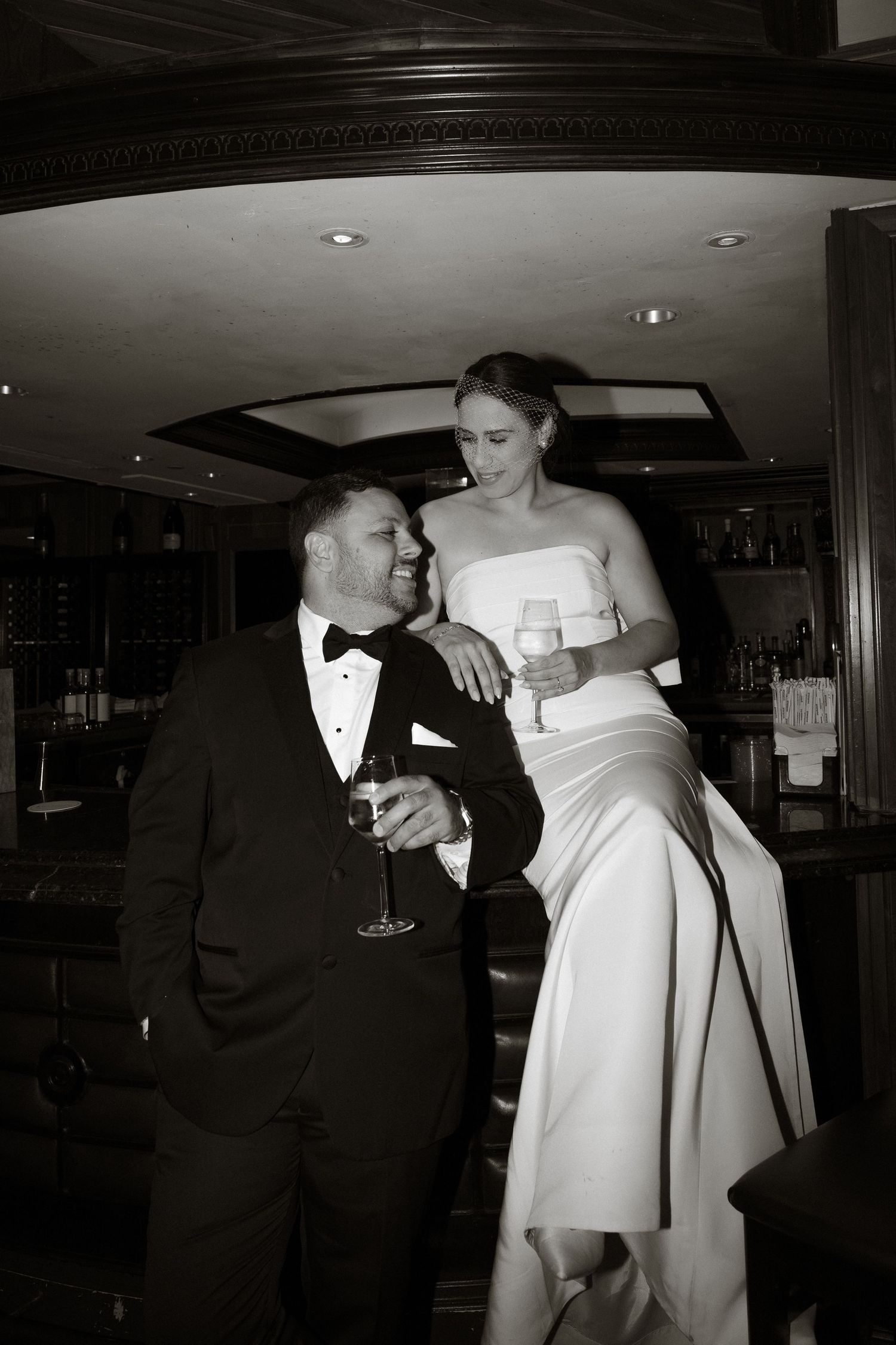 Black and white photo of a couple in formal attire sharing an intimate moment with champagne at an elegant bar.