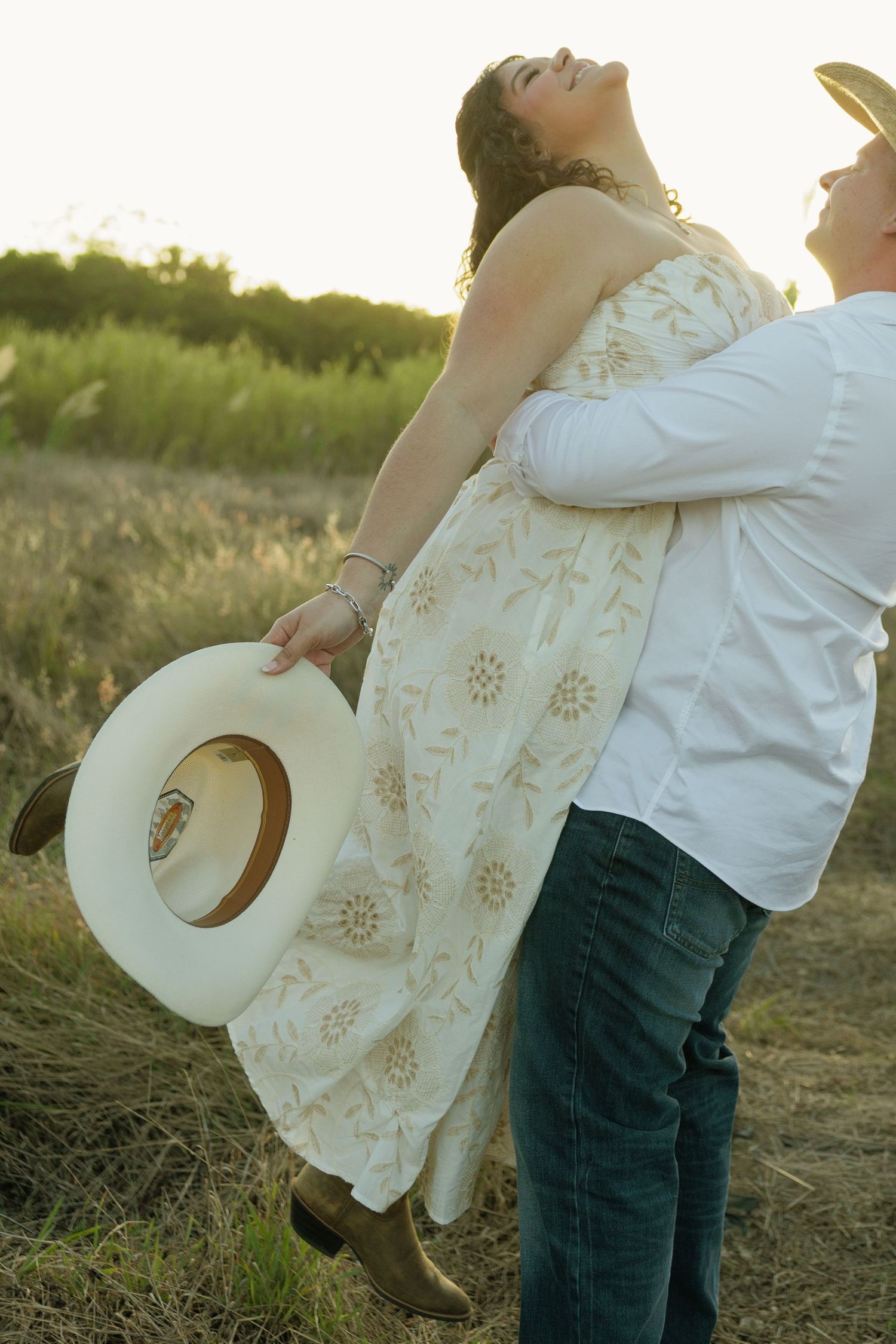 Couple embracing at golden hour in a field, woman in floral dress holding white cowboy hat, man in white shirt and jeans.